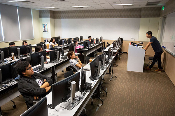 A man stands at a lectern at the front of a classroom with rows of desks lined with computer monitors.