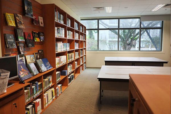 A library with shelves of books and desks. A window looks out to a tree outside.