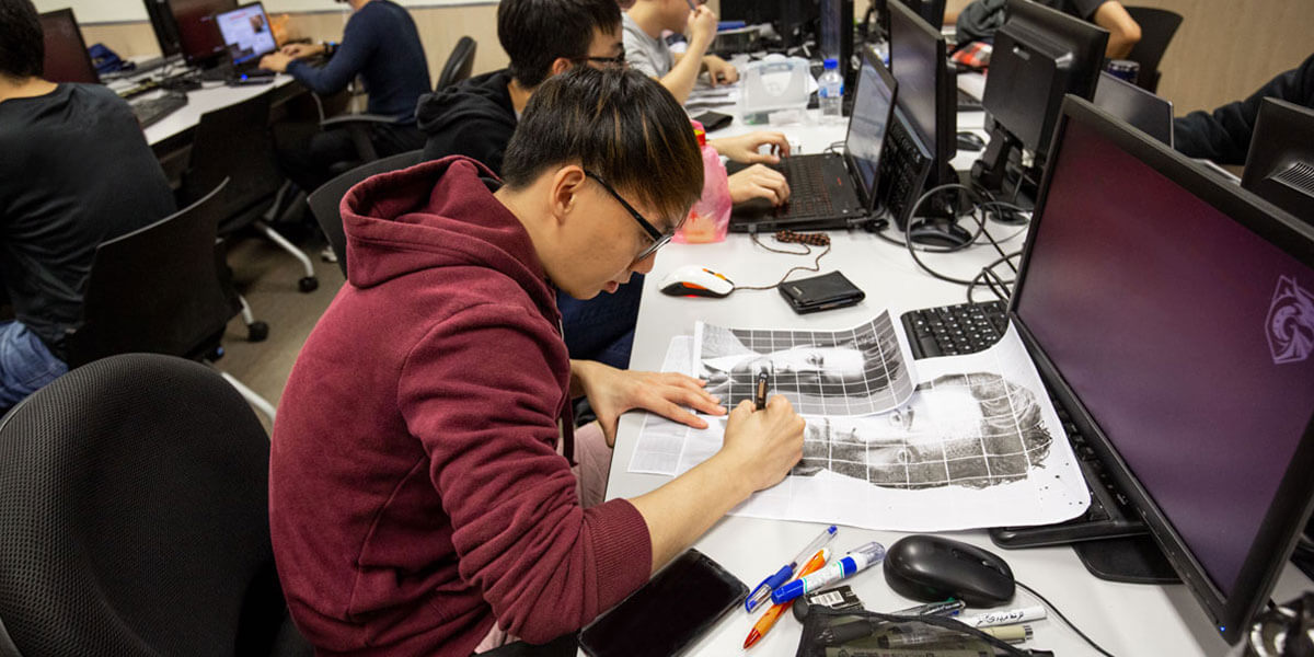 Sitting at a desk, a student in a red sweater sketches a recreation of a tiled, black-and-white image of a man's face.