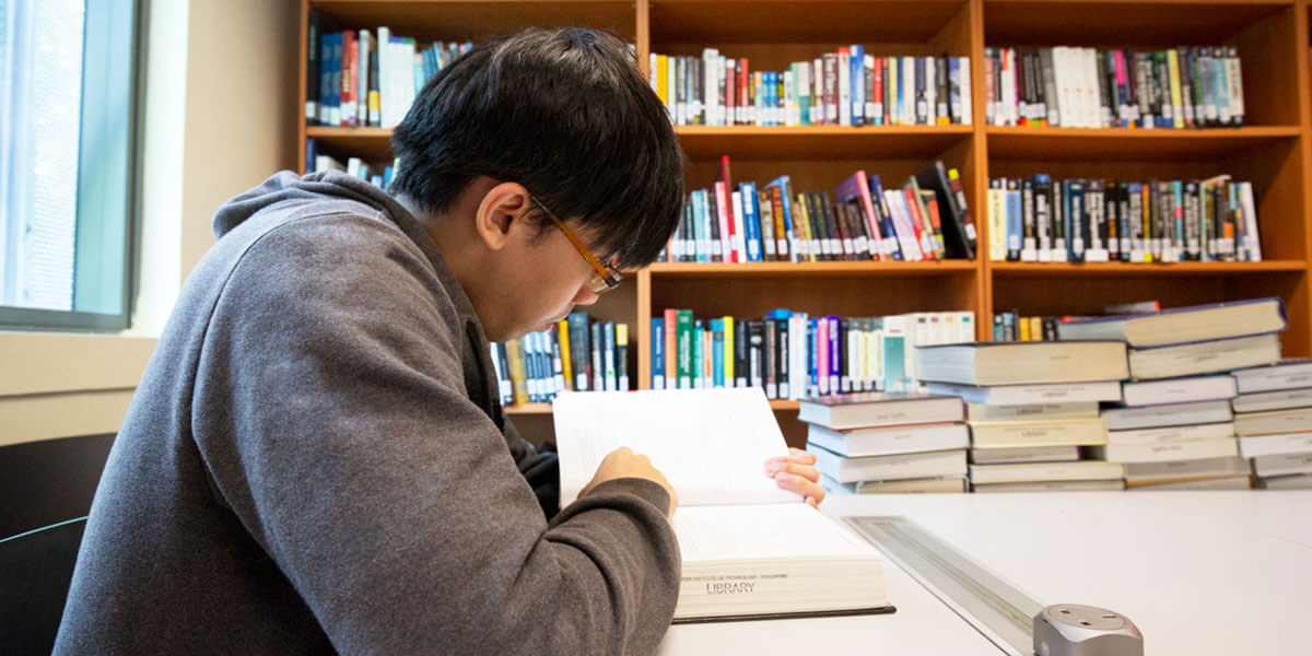 A man reads a thick textbook at an off-white desk with shelves of books seen in the background.