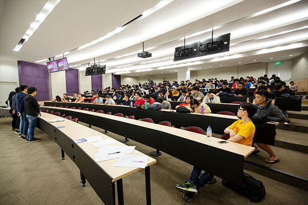 A line of students stands at the front of an academic auditorium with students filling the desks extending toward the back.