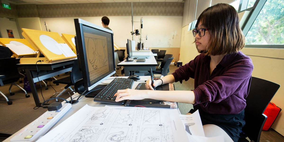 In an animation classroom with multiple light tables, a woman sits at a computer illustrating with a design tablet.