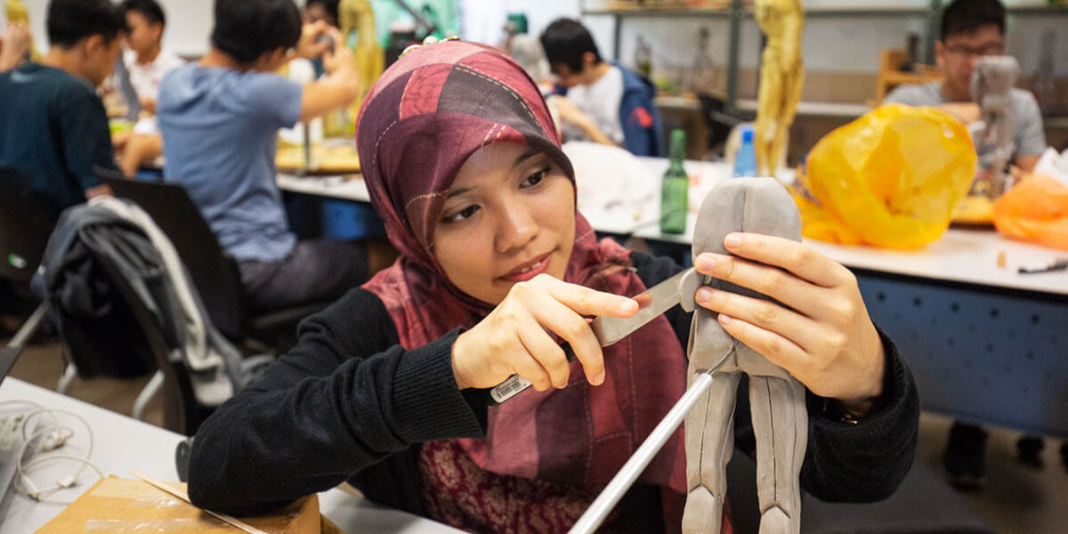 A woman looks intently at the modeling clay in her hands as she uses a metal implement to sculpt a human body.