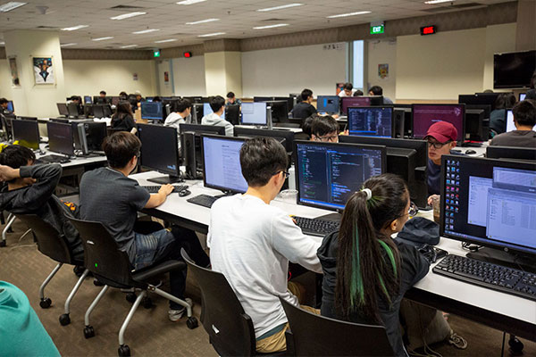 Students sit at rows upon rows of computer monitors and keyboards in a large fluorescent-light-lit computer lab.