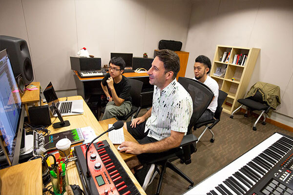 A group of people look at a large computer monitor in a room surrounded by audio speakers and digital piano keyboards.