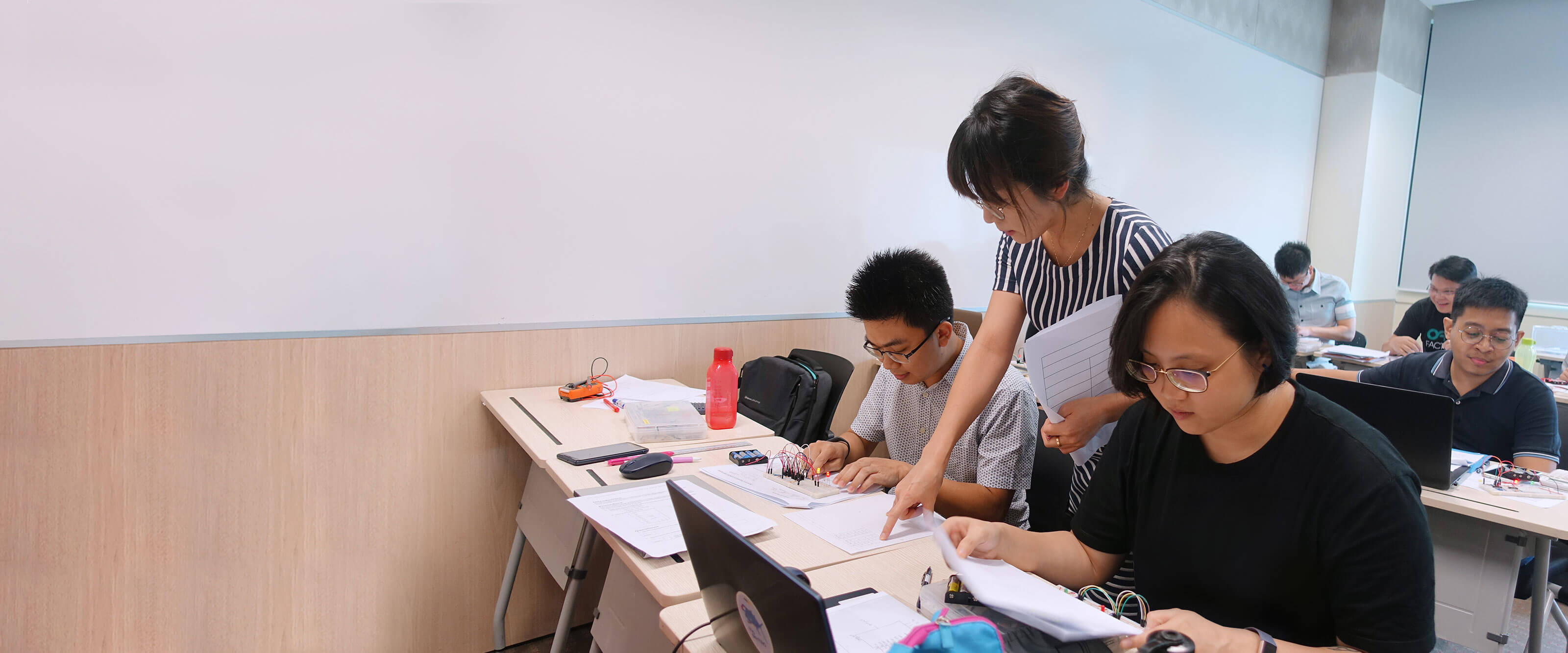 An instructor looks over the shoulder of a seated student who is working on a small electronic circuit board.