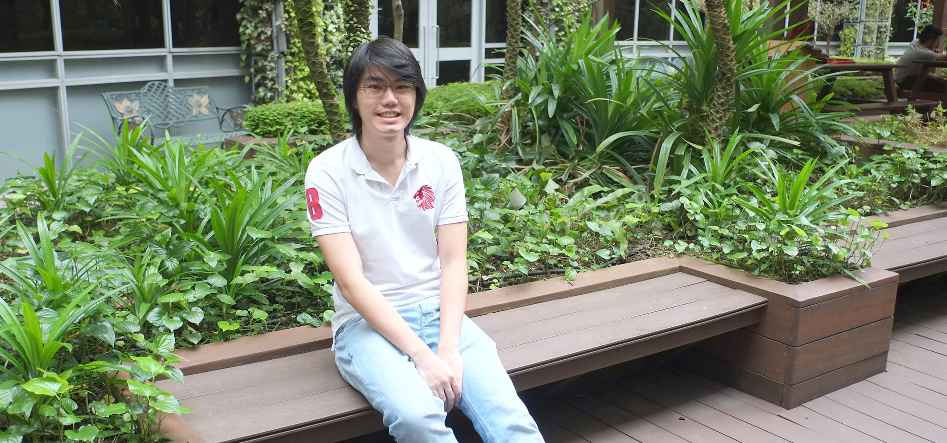DigiPen graduate Cheng Ding Xiang poses for a photo sitting at a bench in an atrium area full of plants and trees