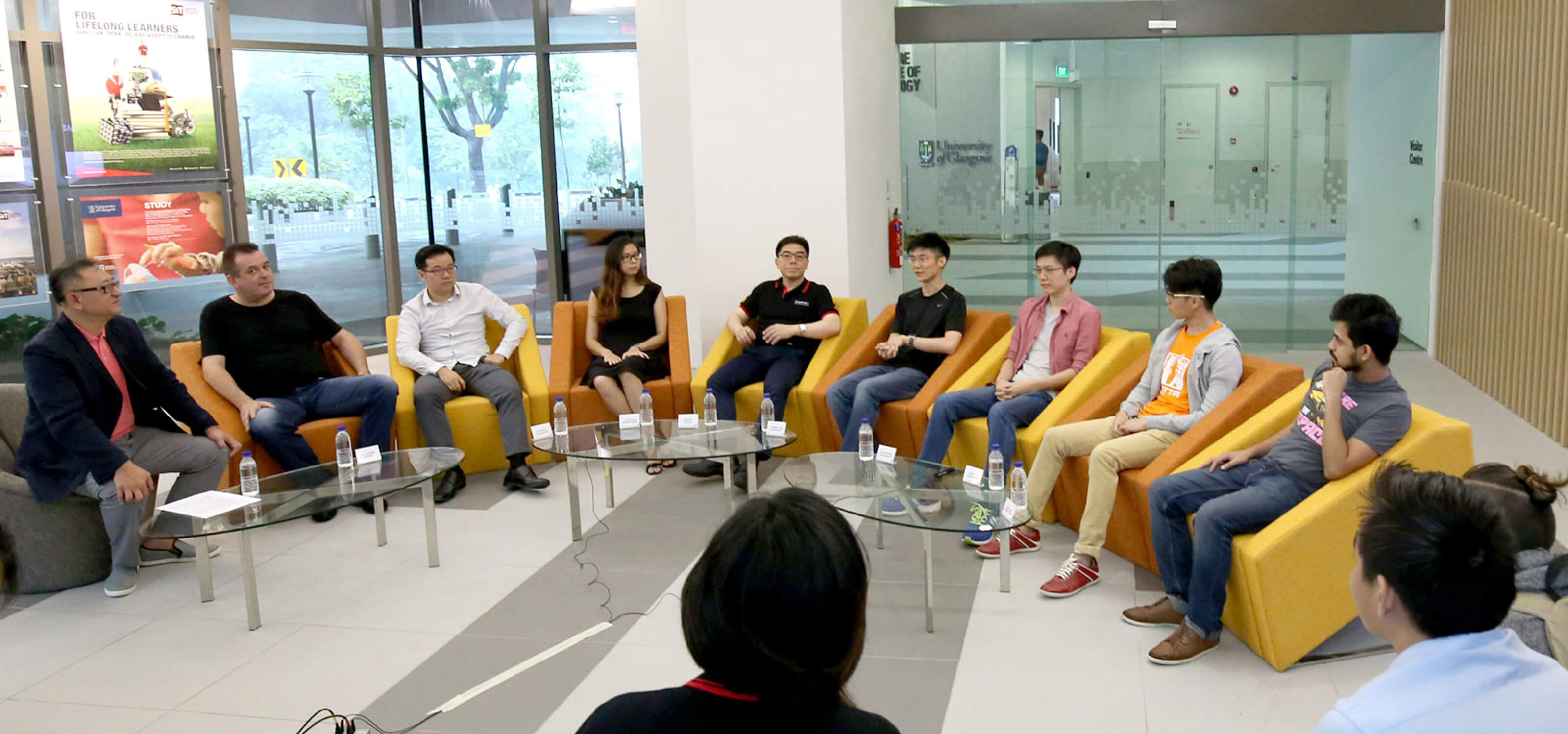 Group of industry professionals sit in orange and yellow chairs in a foyer walled in by glass windows