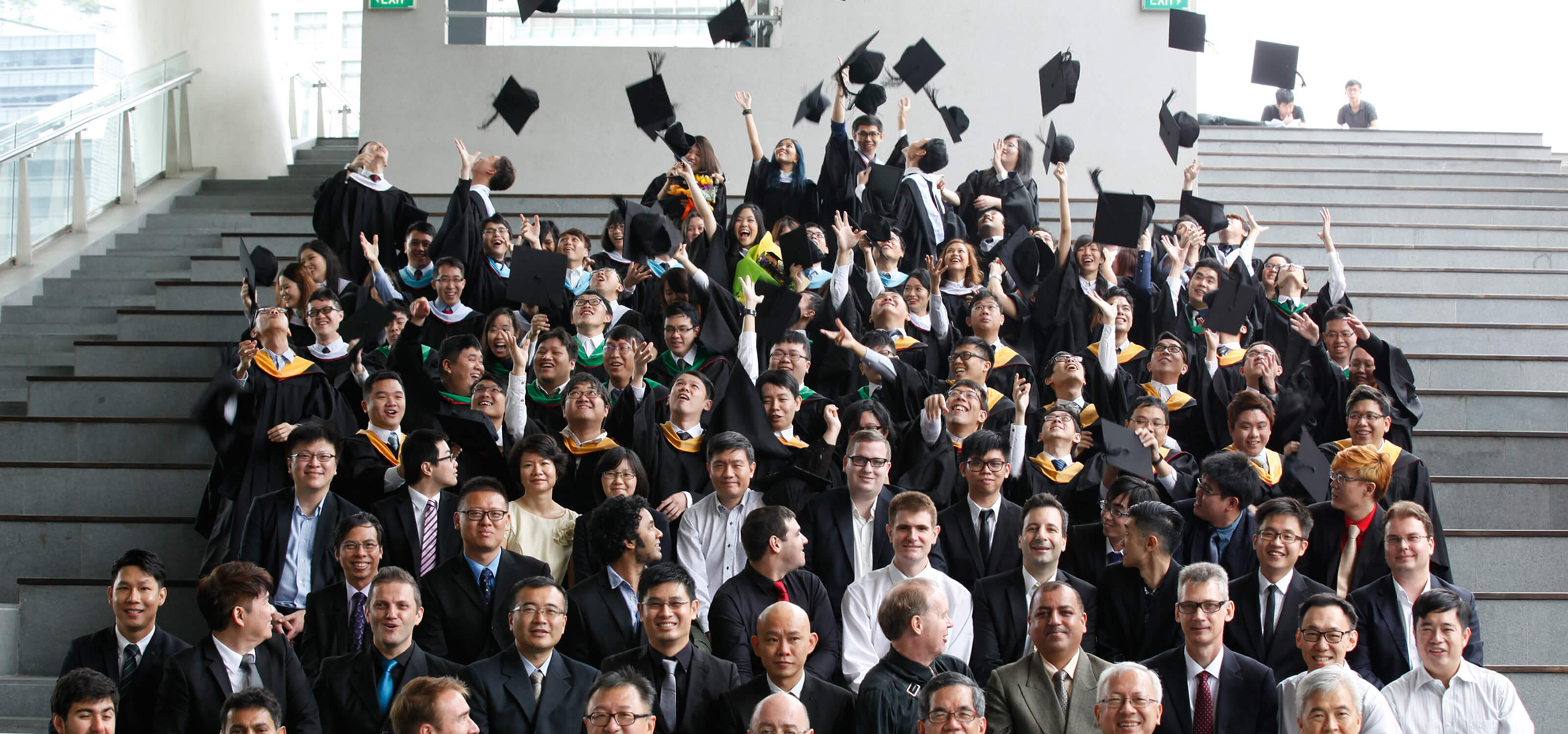 Dozens of newly graduated DigiPen alumni toss their caps in the air as faculty sits below them in bleacher seating