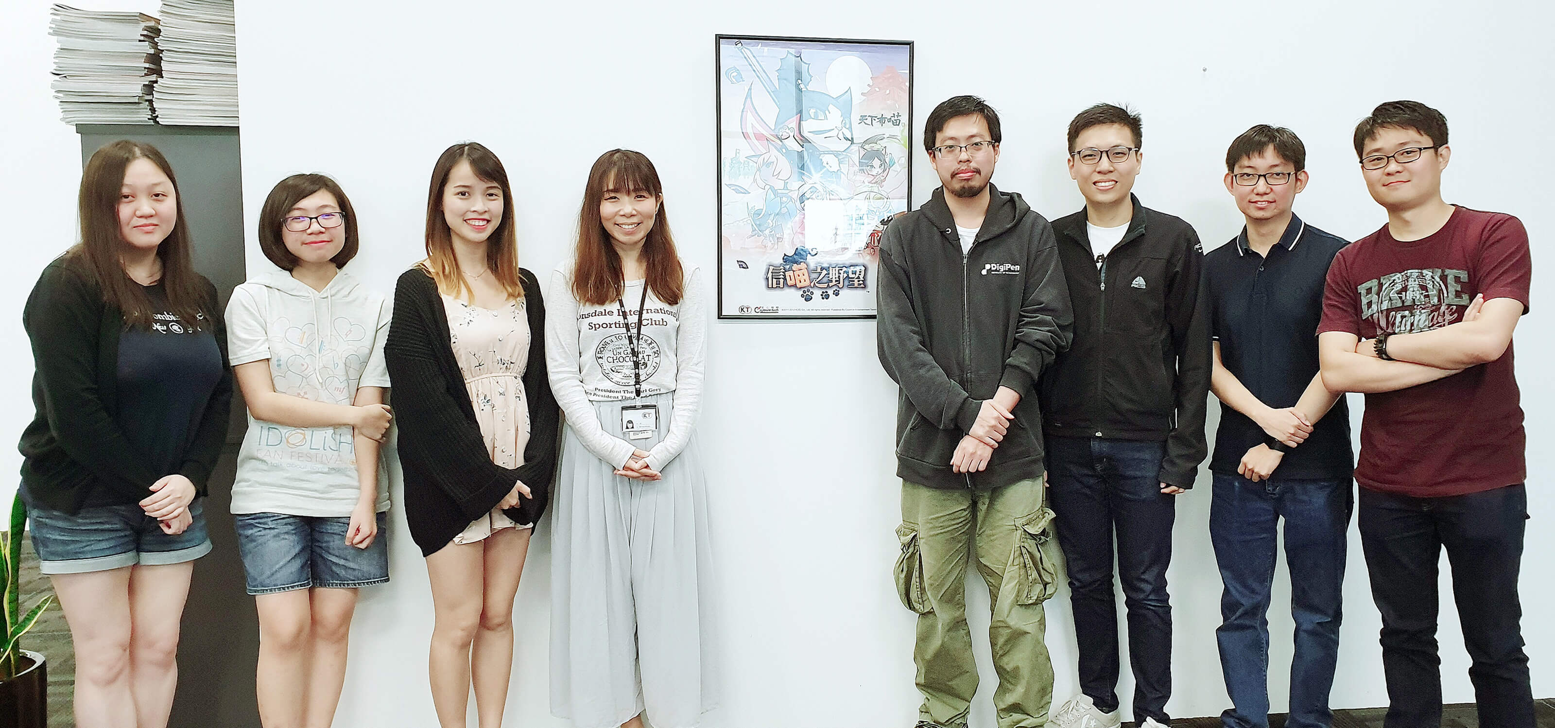 Group photo of the 8 DigiPen (Singapore) alumni who worked on Koei Tecmo's Nyapuri standing in office by a poster of the game.