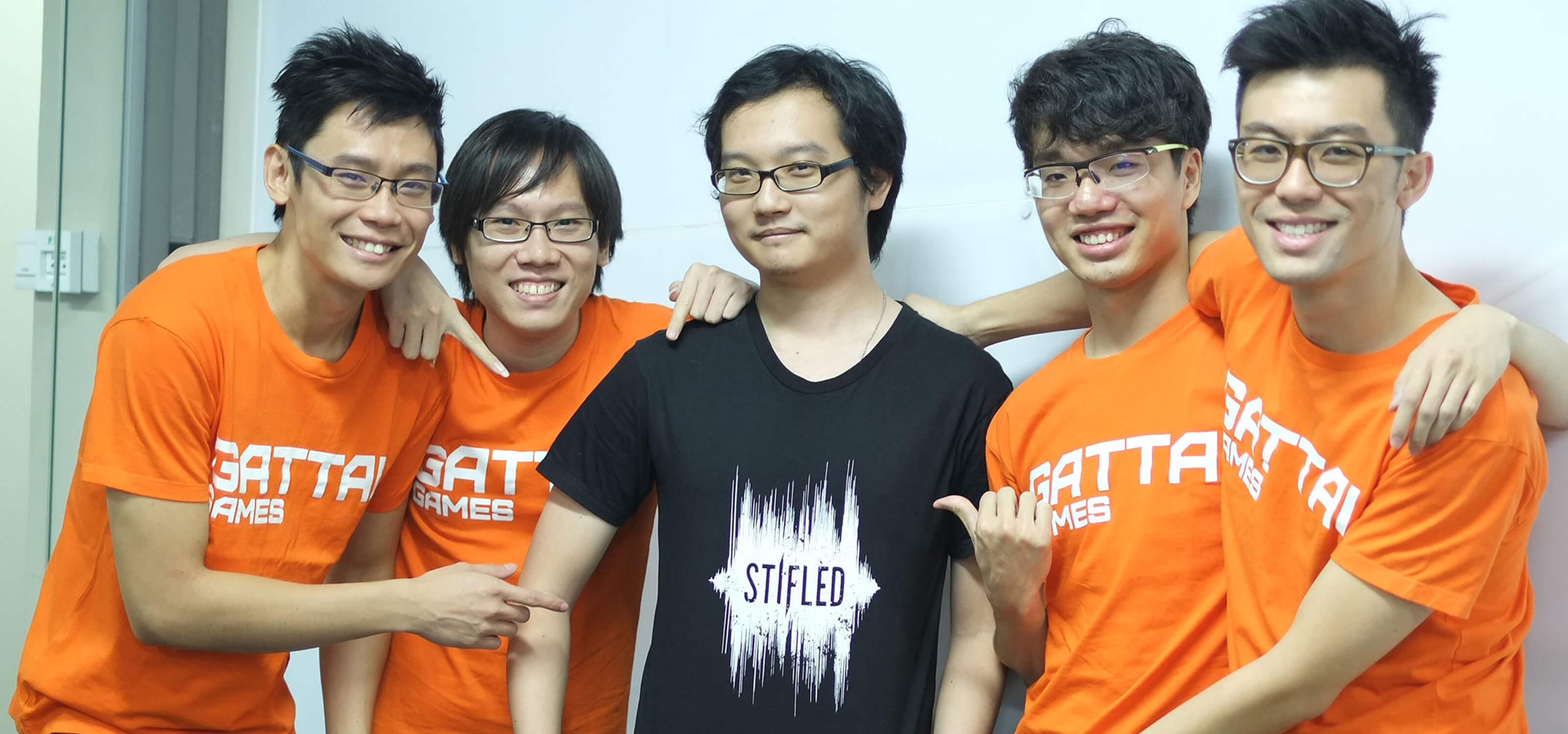 Five members of the Stifled dev team, four in orange t-shirts reading "Gattai Games," and one in a black t-shirt