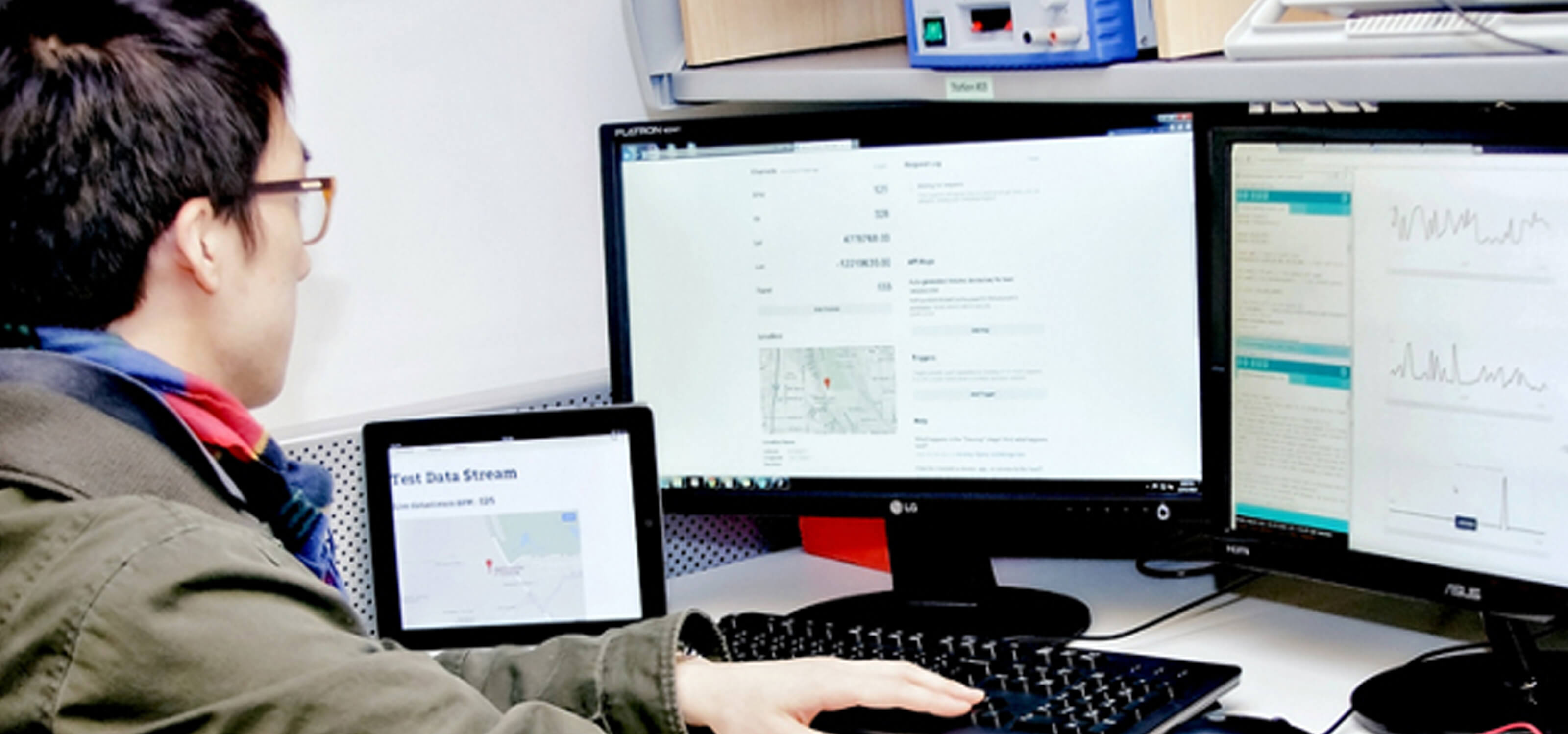 Man sits at a desk looking at two computer monitors. A tablet computer and electrical wiring are also on the desk