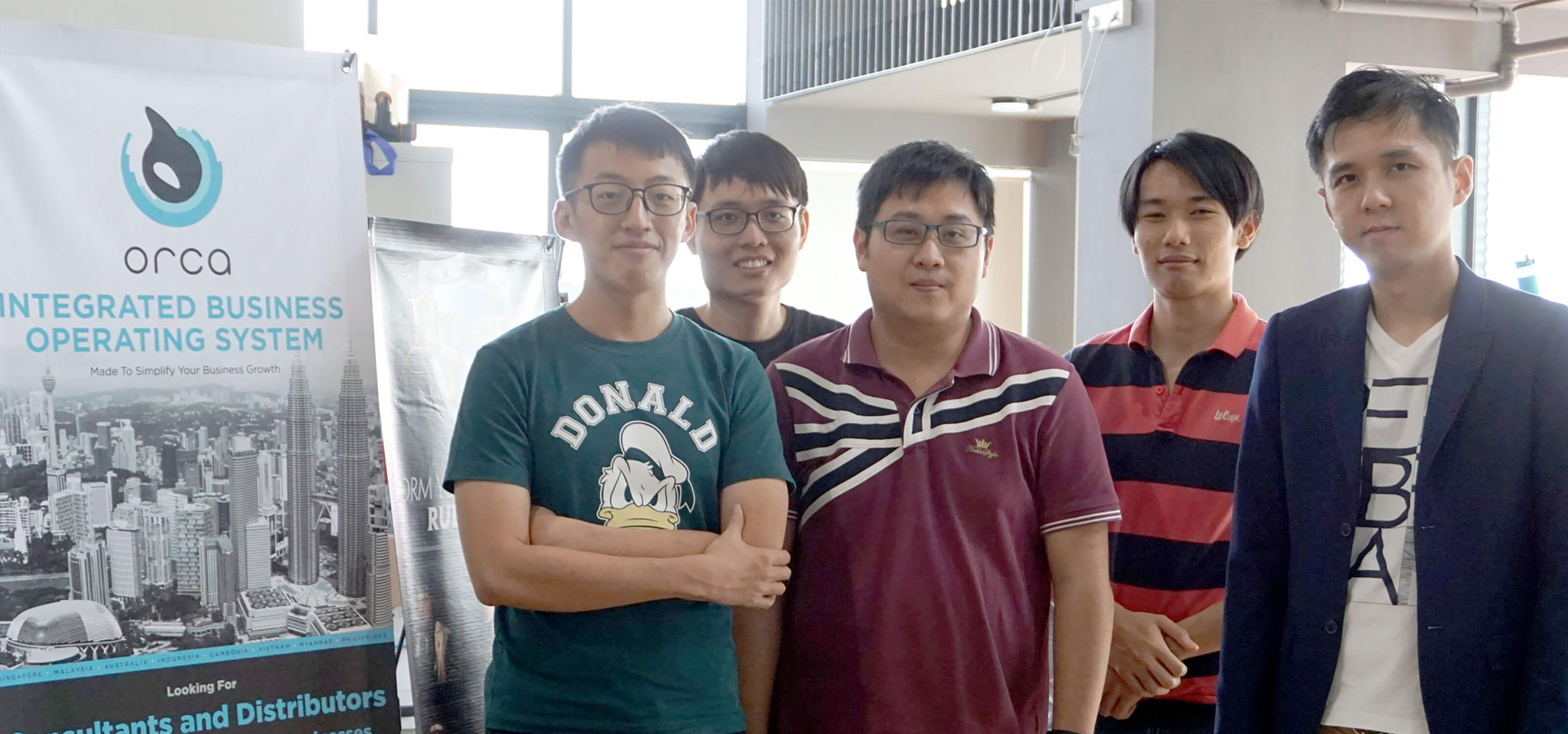A group of DigiPen (Singapore) alumni pose next to a banner advertising the ORCA Integrated Business Operating System