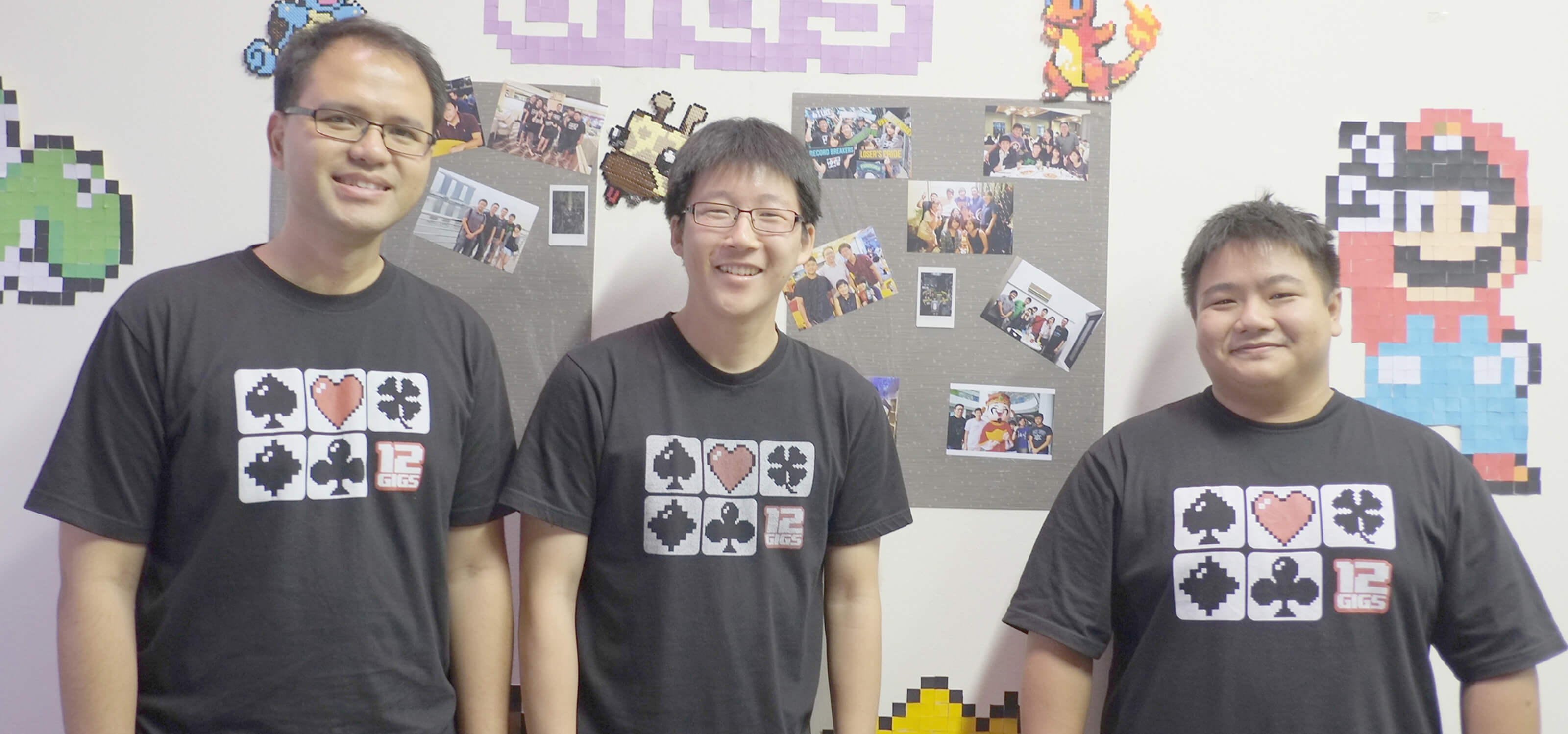 Three DigiPen graduates stand in front of a wall decorated with pixel art and the 12 Gigs logo wearing black t-shirts