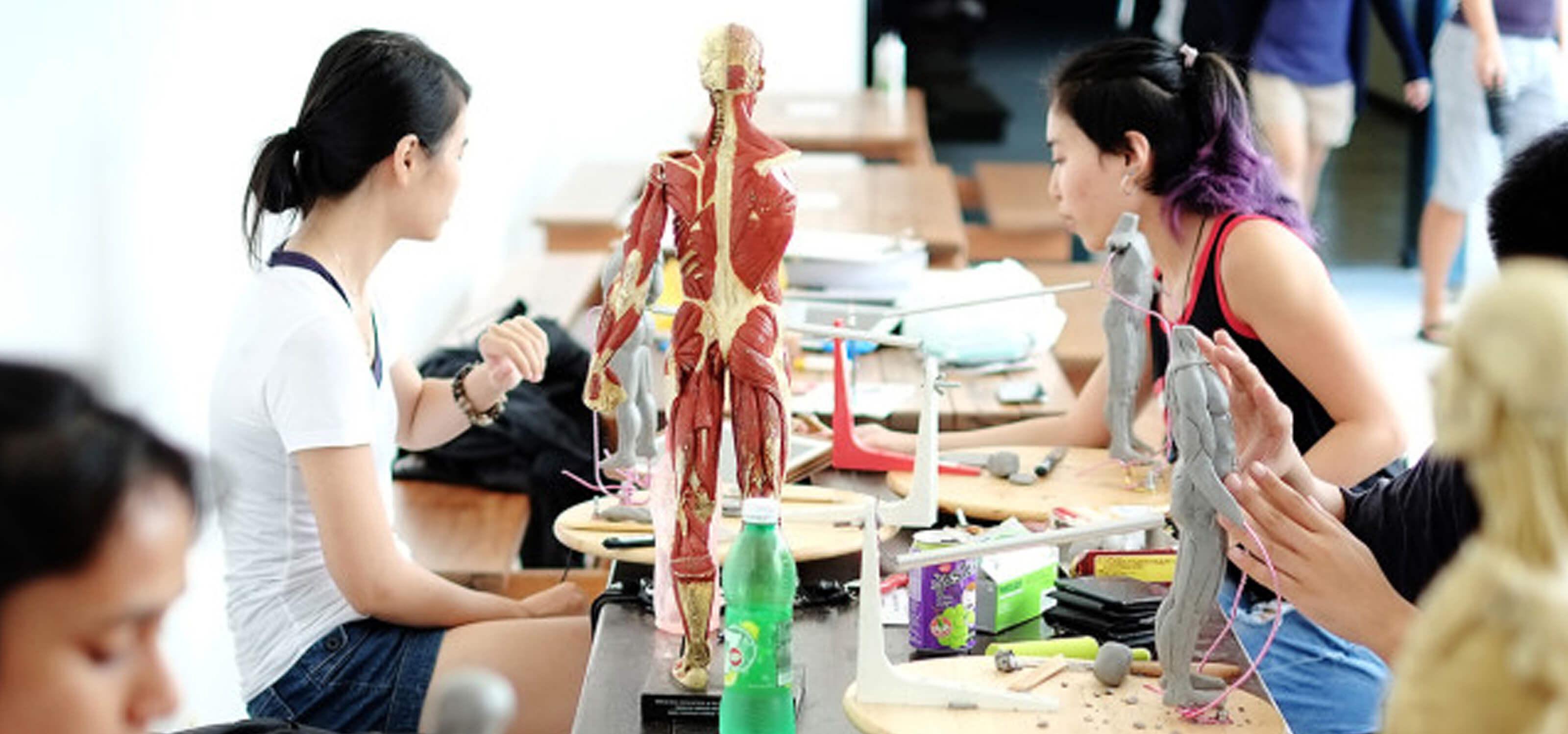 Students seated at a picnic table sculpt human figures out of clay. A plastic human musculoskeletal figure acts as a model.