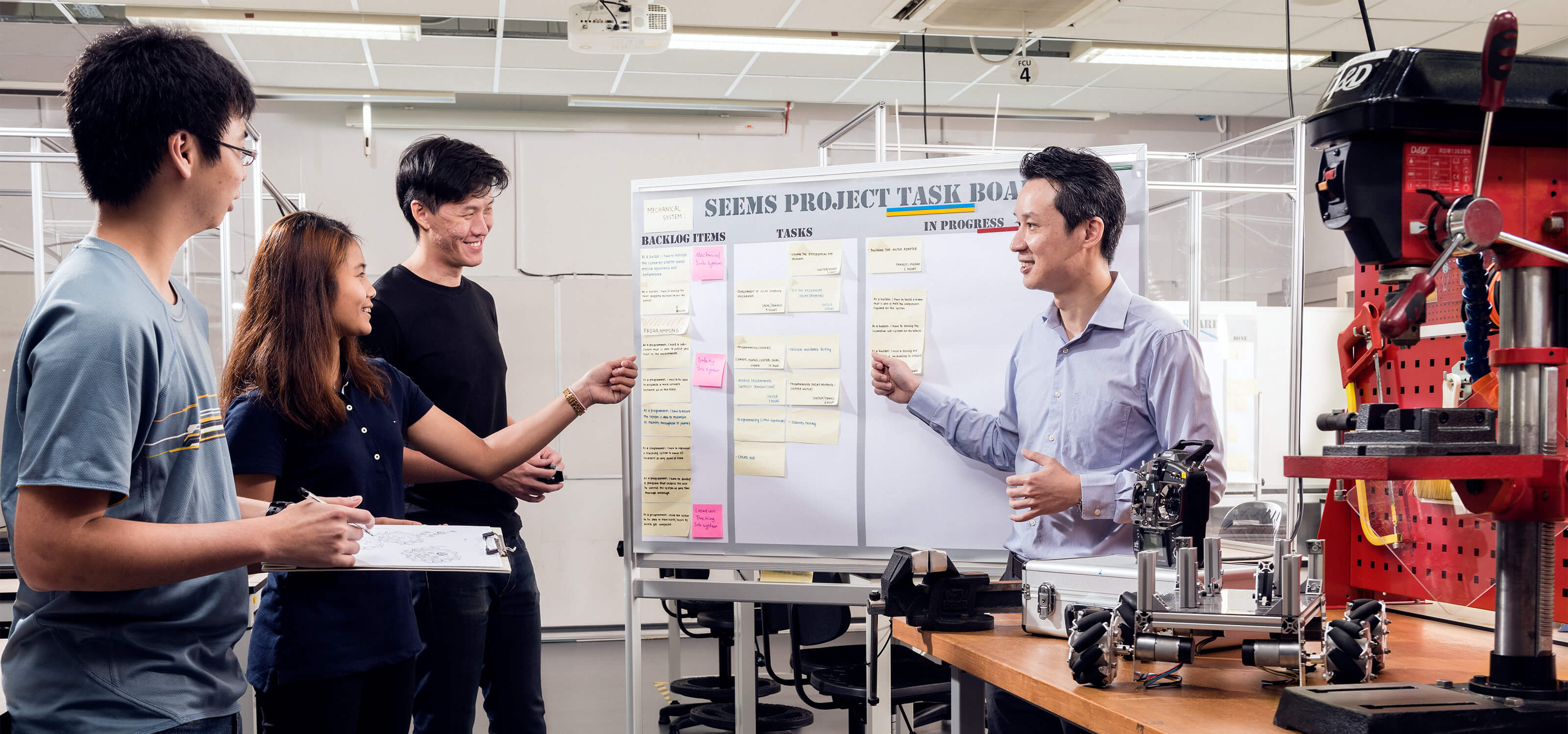 Three students and a teacher stand at a task board in a classroom lab.