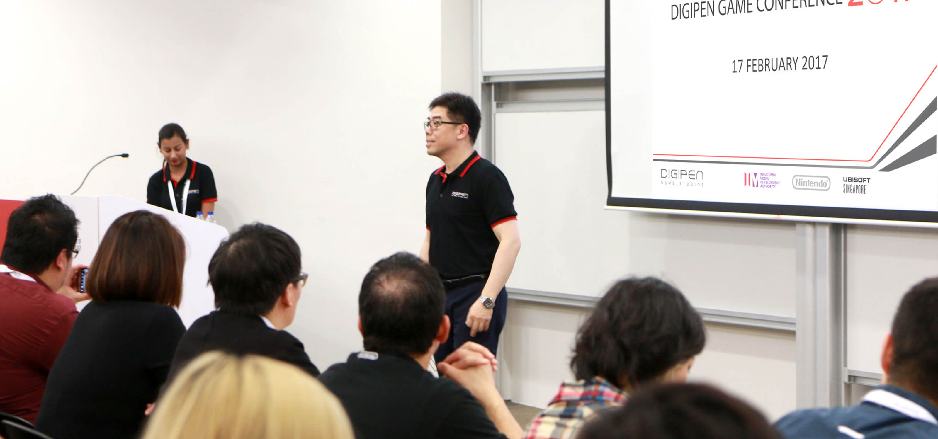 DigiPen COO Jason Chu addresses a room of industry professionals in a conference room. A projection screen is behind him.