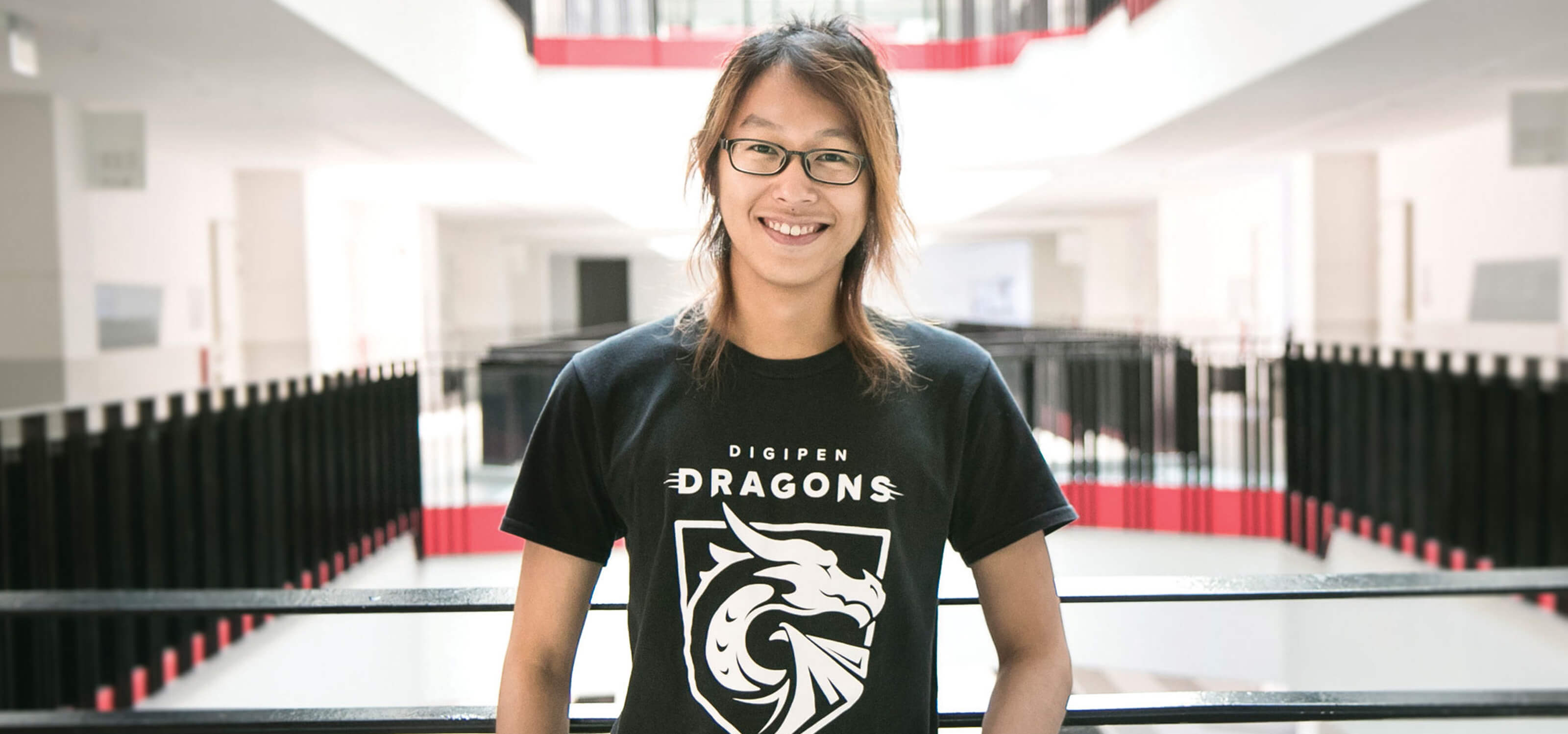 DigiPen graduate Jovi Kartolo poses for a photo at a railing on the second floor wearing a black DigiPen Dragons t-shirt