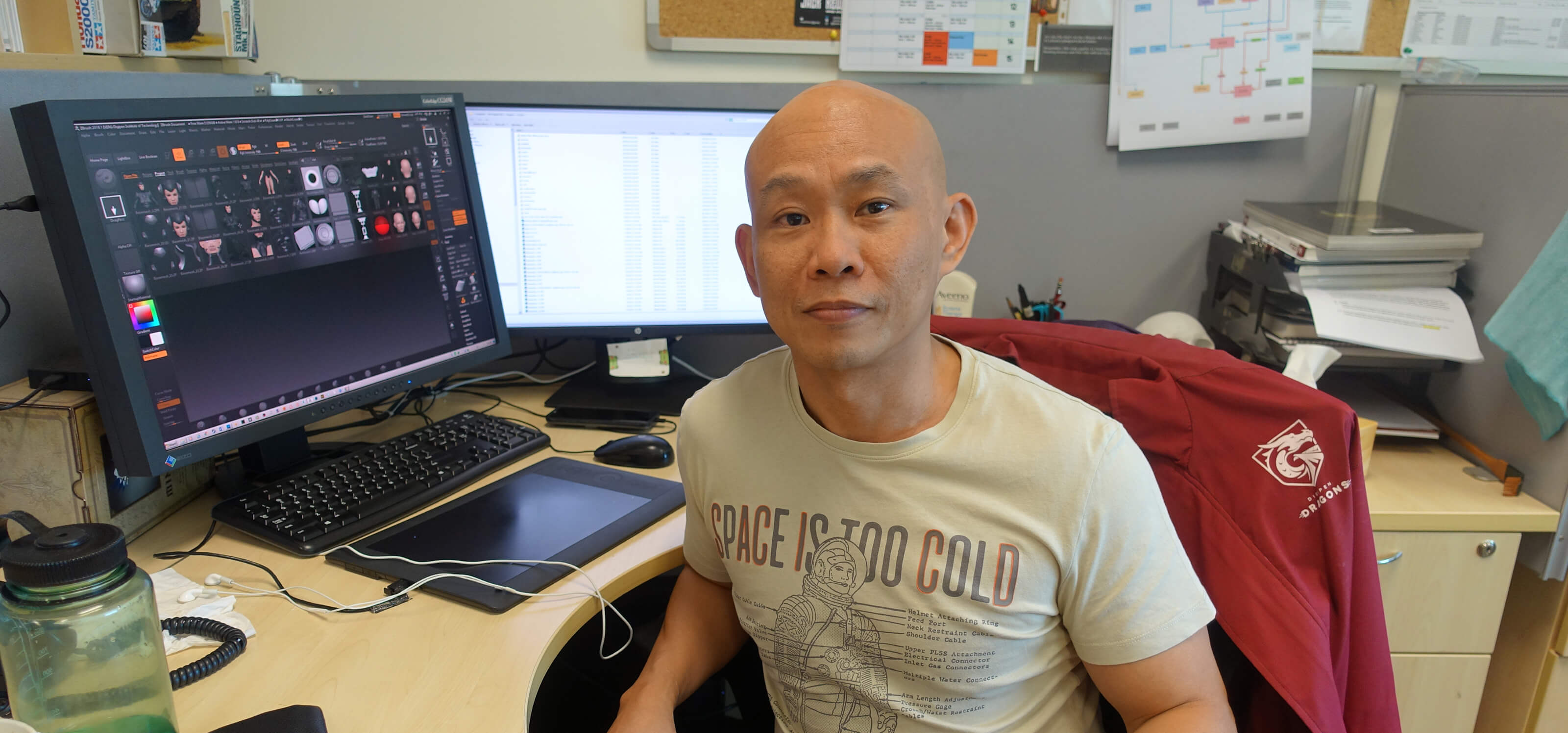 Professor Marc Tan, wearing a tan T-shirt, faces the camera while sitting at his office desk; computer monitor shows thumbnails of his 3D artwork.