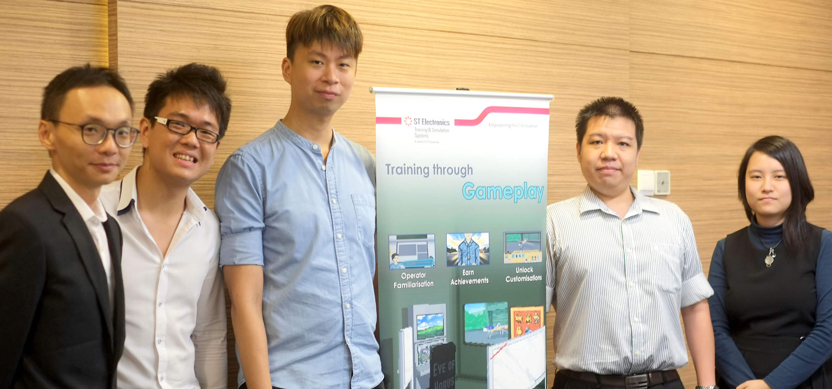 Team from ST Electronics poses for a photo with a vertical banner explaining a learning initiative using gameplay mechanics