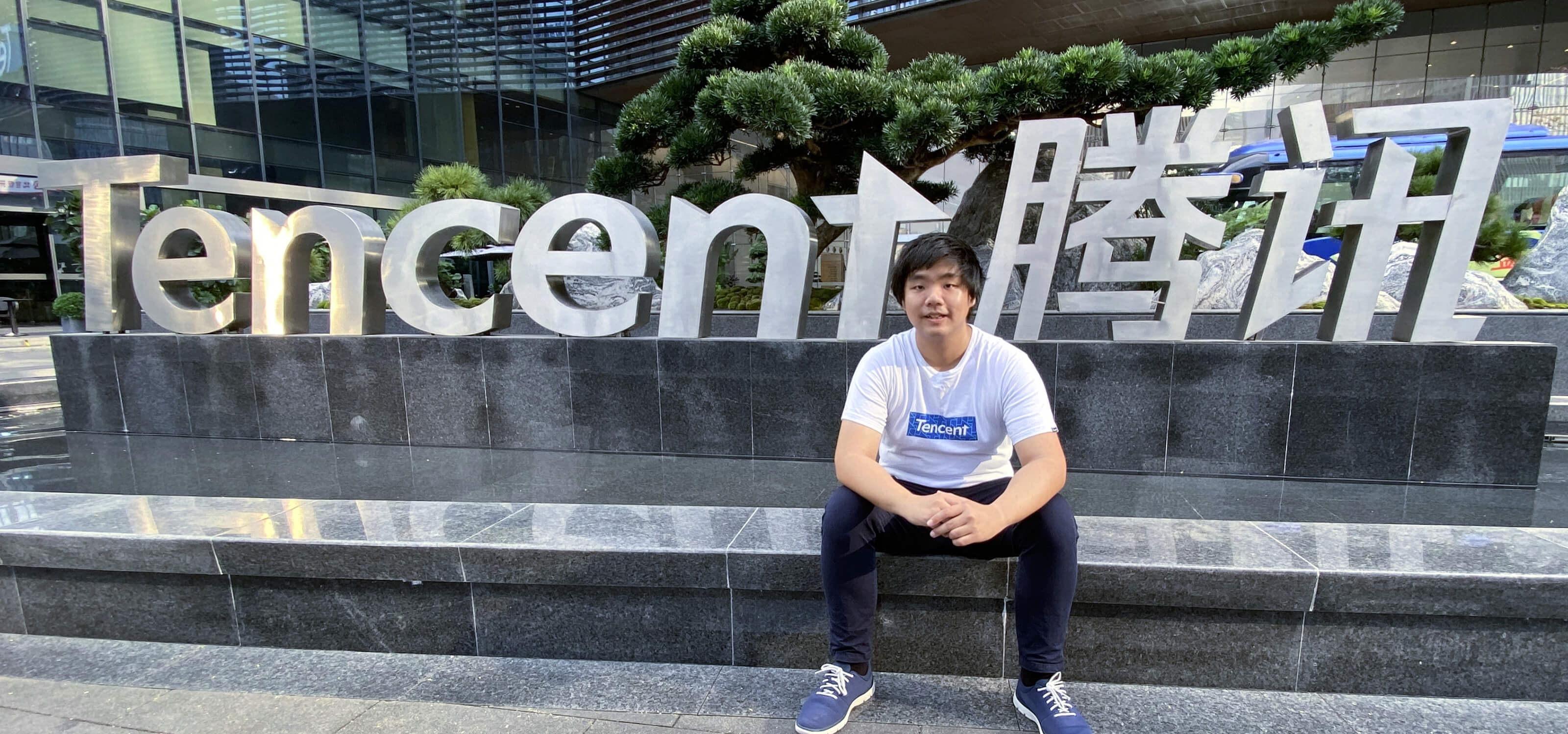 Sim Yi Cong sits by a pool in front of a large Tencent logo sign outside the Tencent headquarters building.