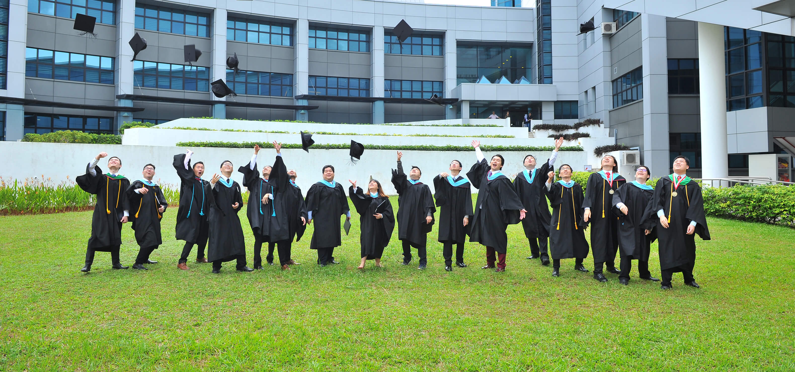Several lined-up graduates throw their graduation caps into the air while standing outside on the grass.