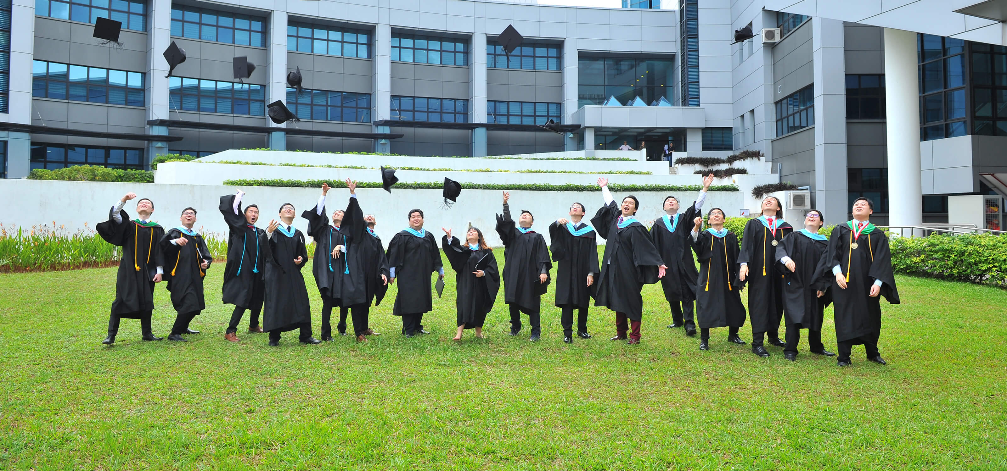 Several lined-up graduates throw their graduation caps into the air while standing outside on the grass.