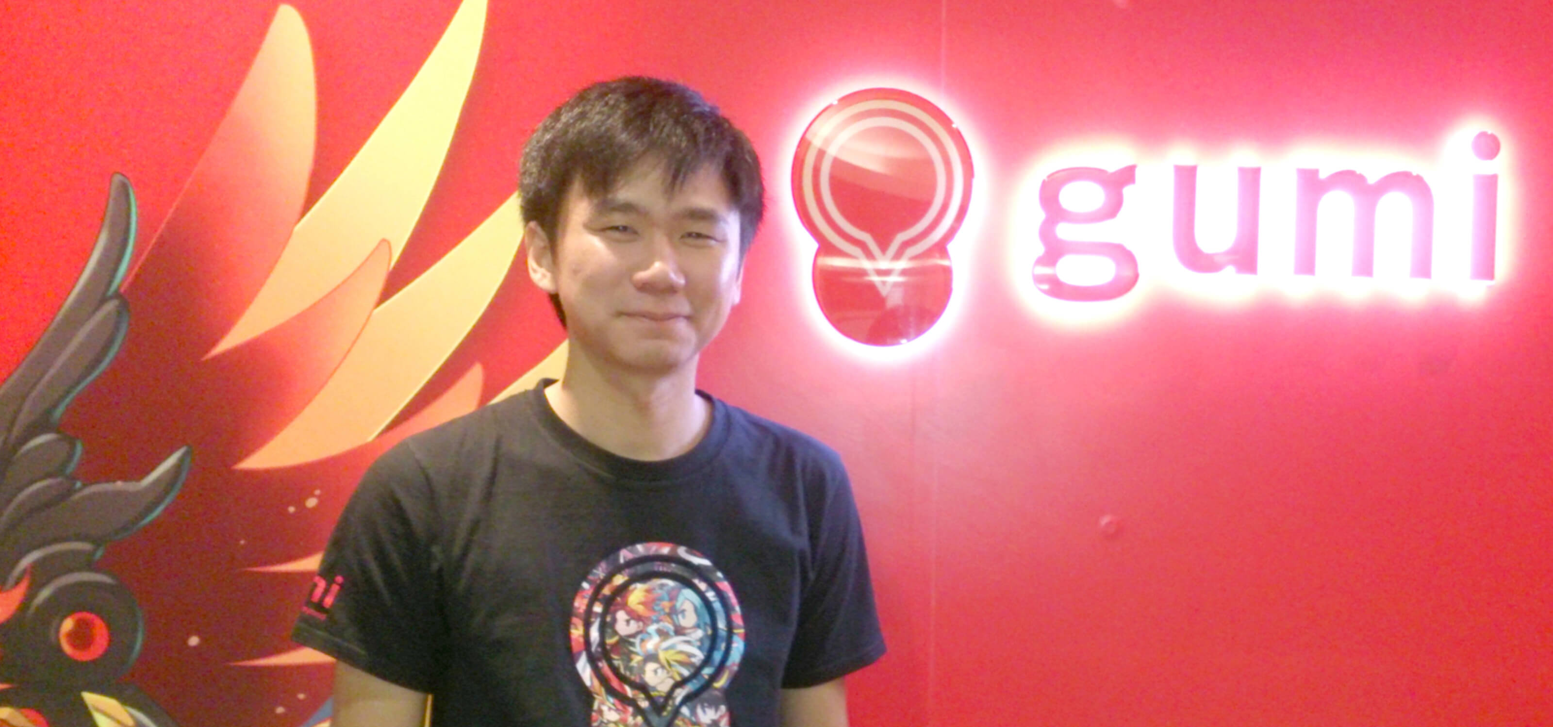 DigiPen graduate Soh Huajin stands in front of a red wall with a glowing logo for gumi Asia behind him