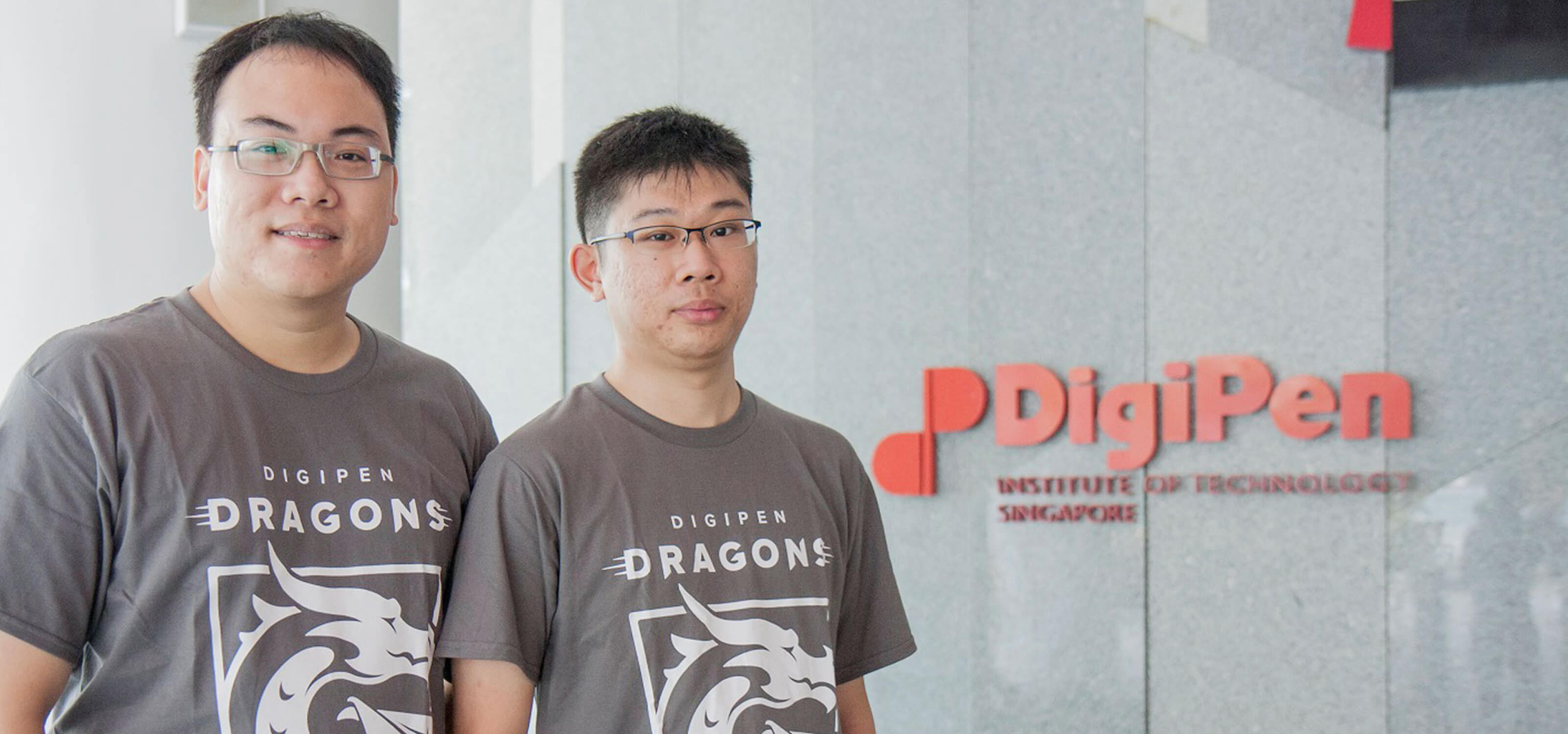 Graduates Tan Tien Wei Keith and Yap Teng Hong pose for a photo in gray DigiPen Dragons t-shirts next to the DigiPen building