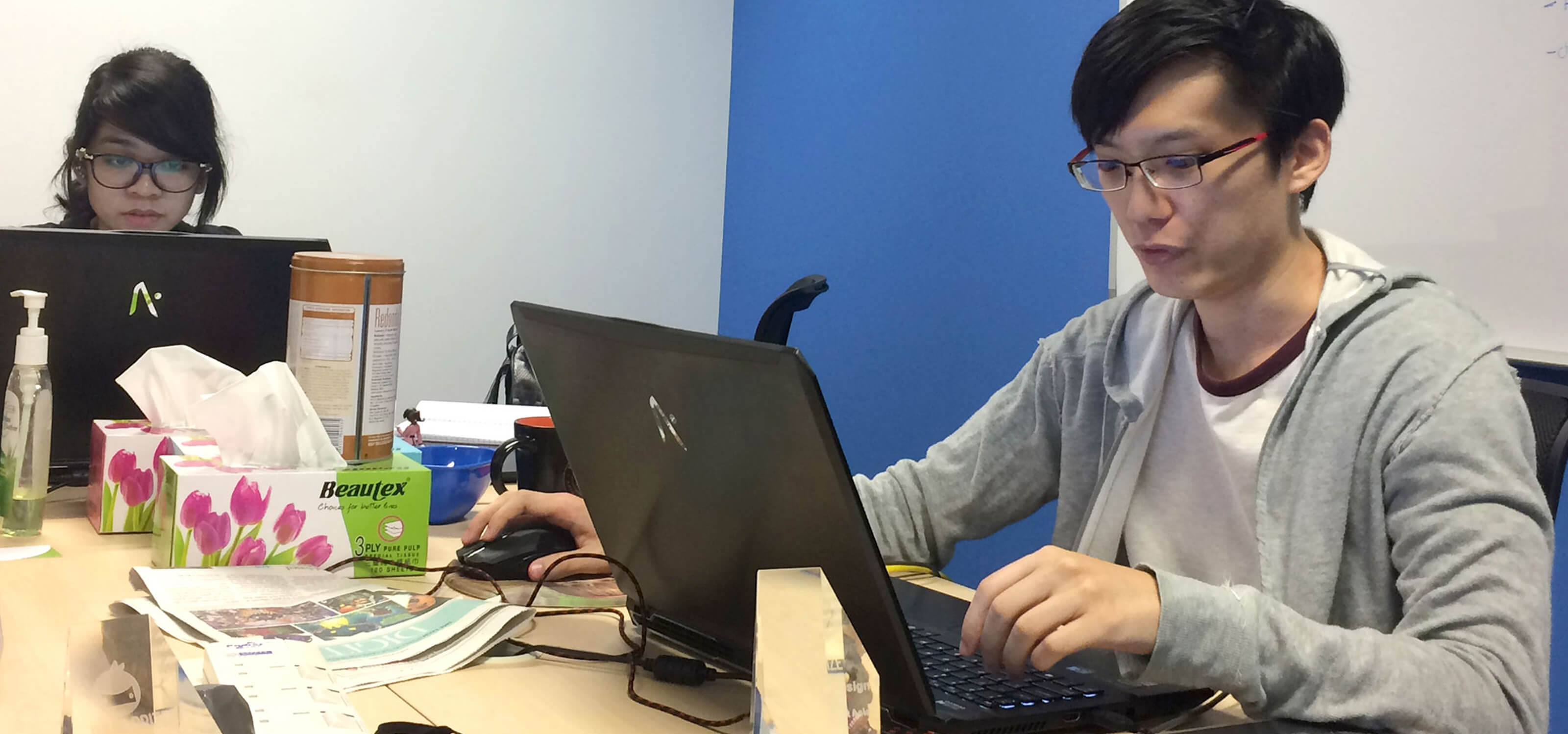 Alumnus Desmond Wong sits at a desk with a laptop computer surrounded by trinkets and awards