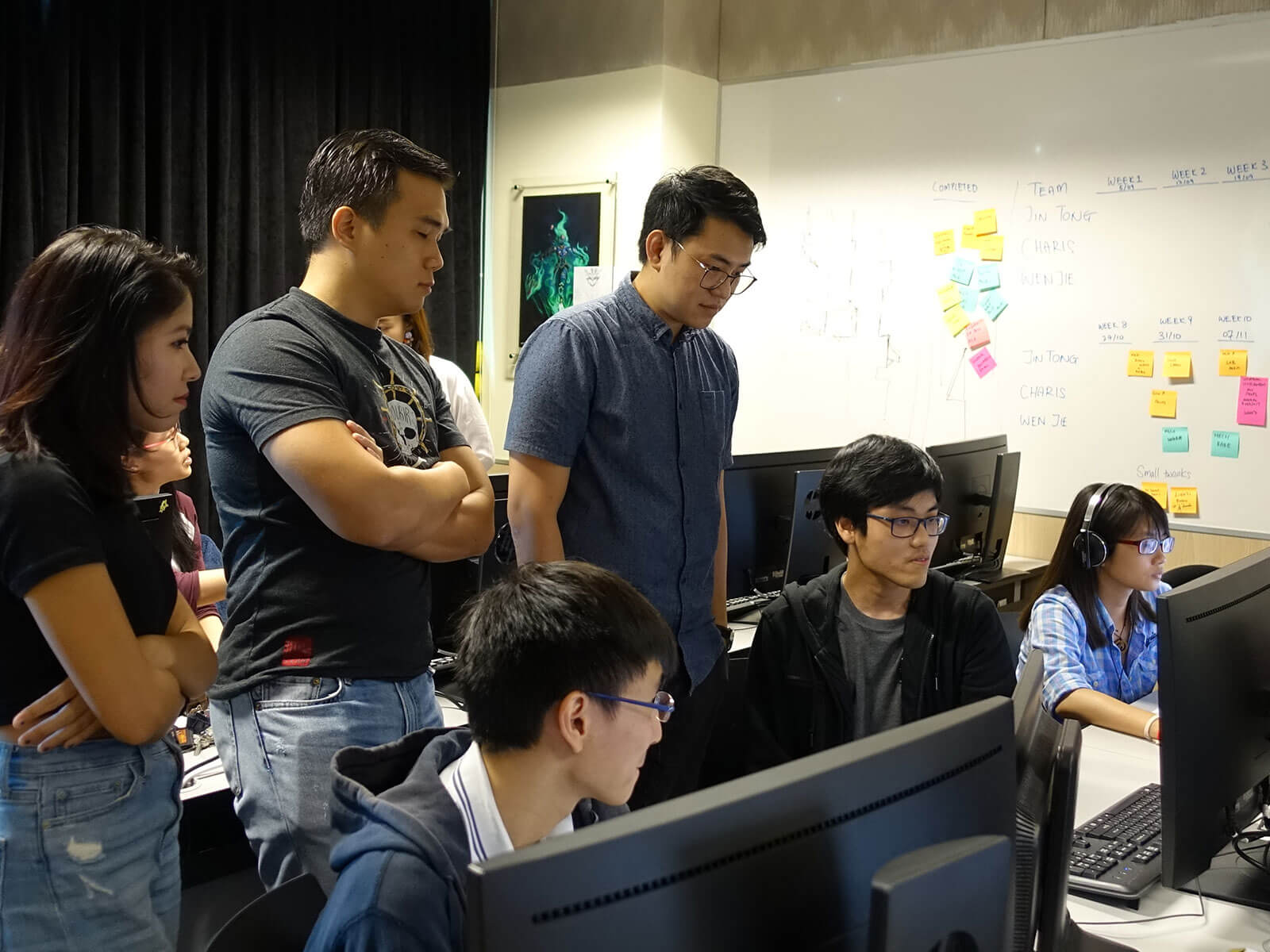 Kobe Sek from Ubisoft Singapore and others look down at a student’s monitor in a computer lab classroom, with students seated at two rows of workstations.