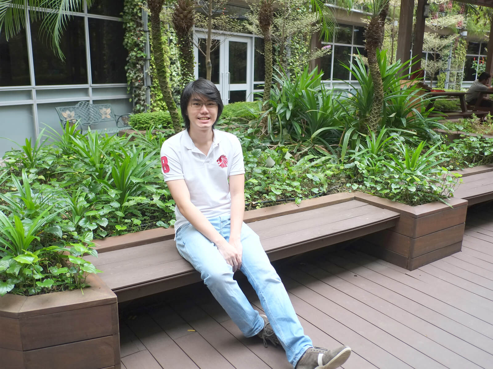 DigiPen graduate Cheng Ding Xiang poses for a photo sitting at a bench in an atrium area full of plants and trees