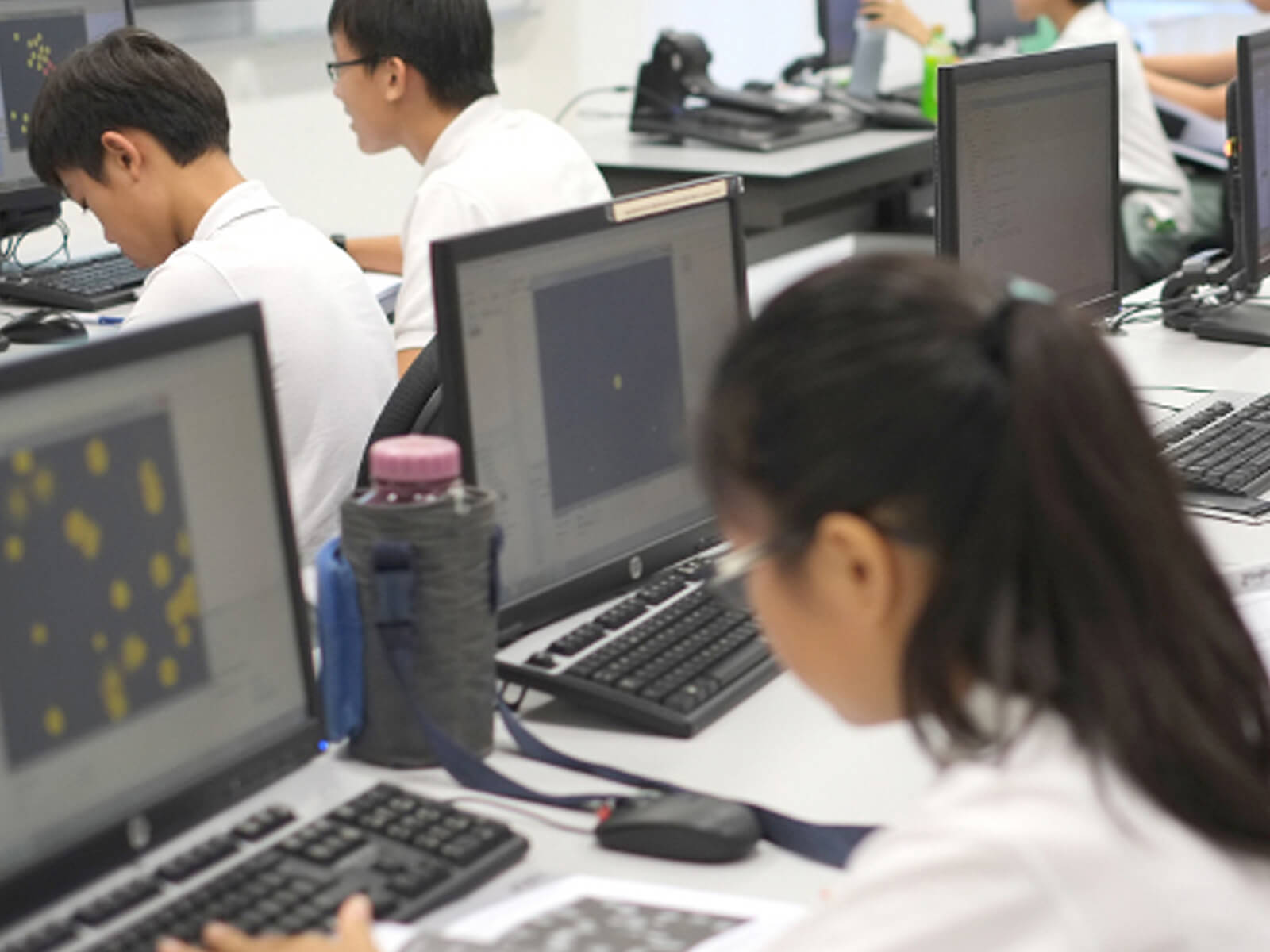 Students seen from over the shoulder, each sitting at a desk with a computer, working on different projects