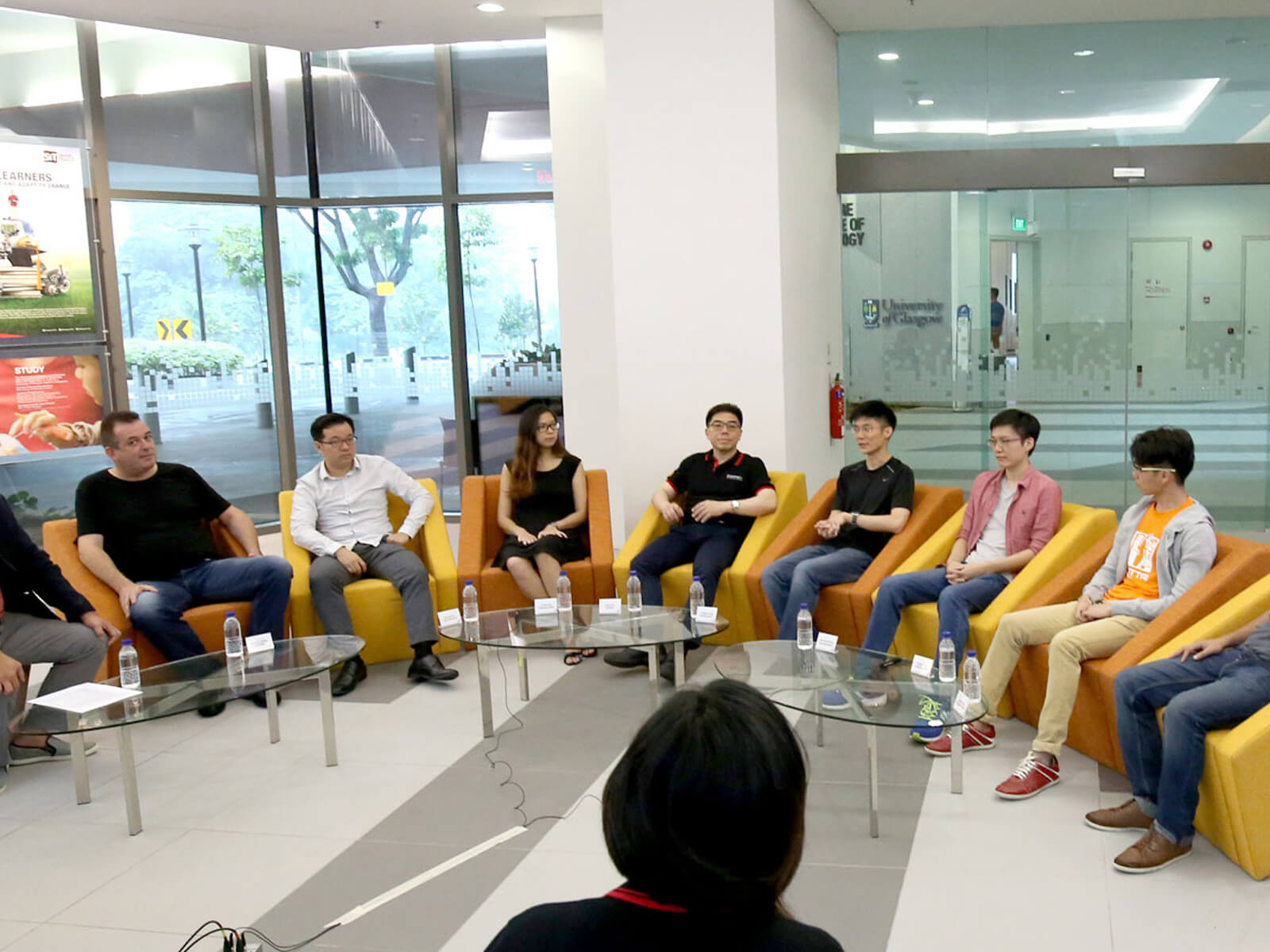 Group of industry professionals sit in orange and yellow chairs in a foyer walled in by glass windows