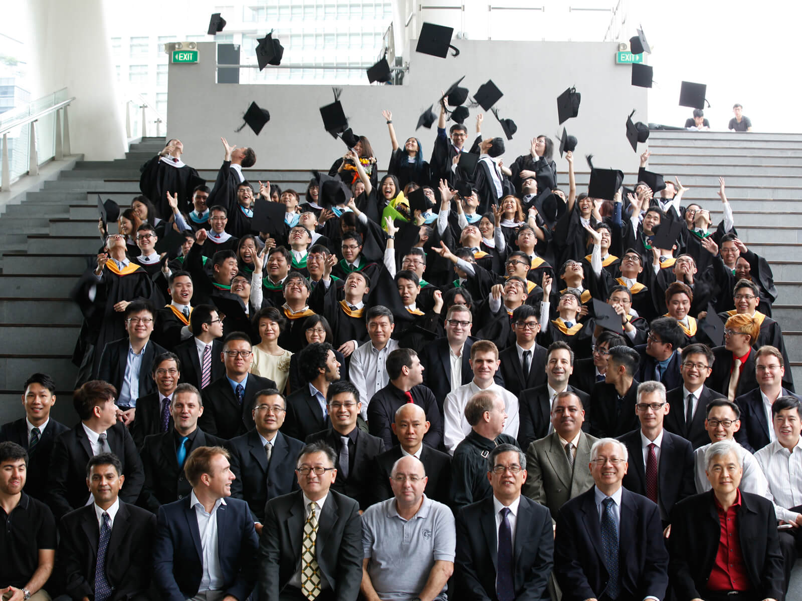 Dozens of newly graduated DigiPen alumni toss their caps in the air as faculty sits below them in bleacher seating