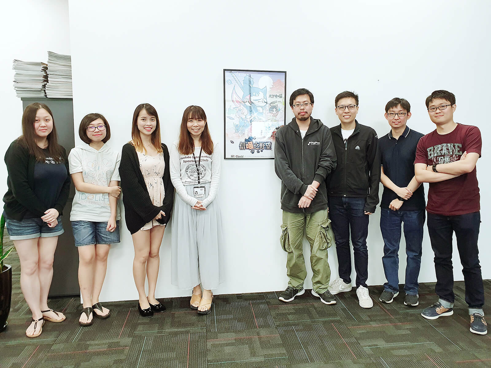 Group photo of the 8 DigiPen (Singapore) alumni who worked on Koei Tecmo's Nyapuri standing in office by a poster of the game.