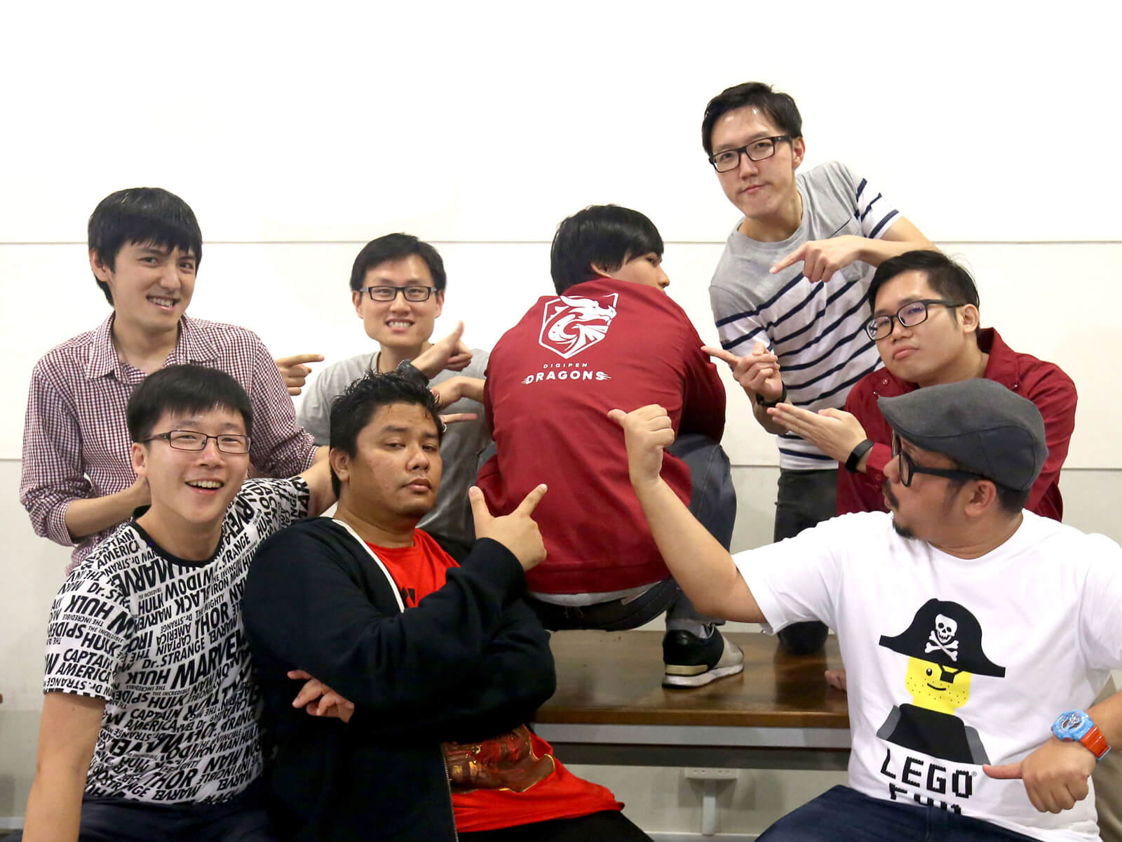 Members of the alumni committee point to the back of another member squatting on a desk who is wearing a red DigiPen shirt