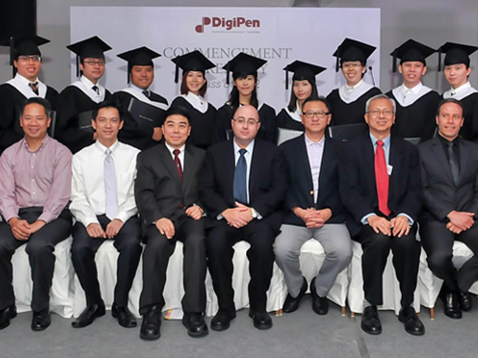 12 DigiPen (Singapore) graduates of the inaugural class pose in cap and gowns behind seated faculty and administration