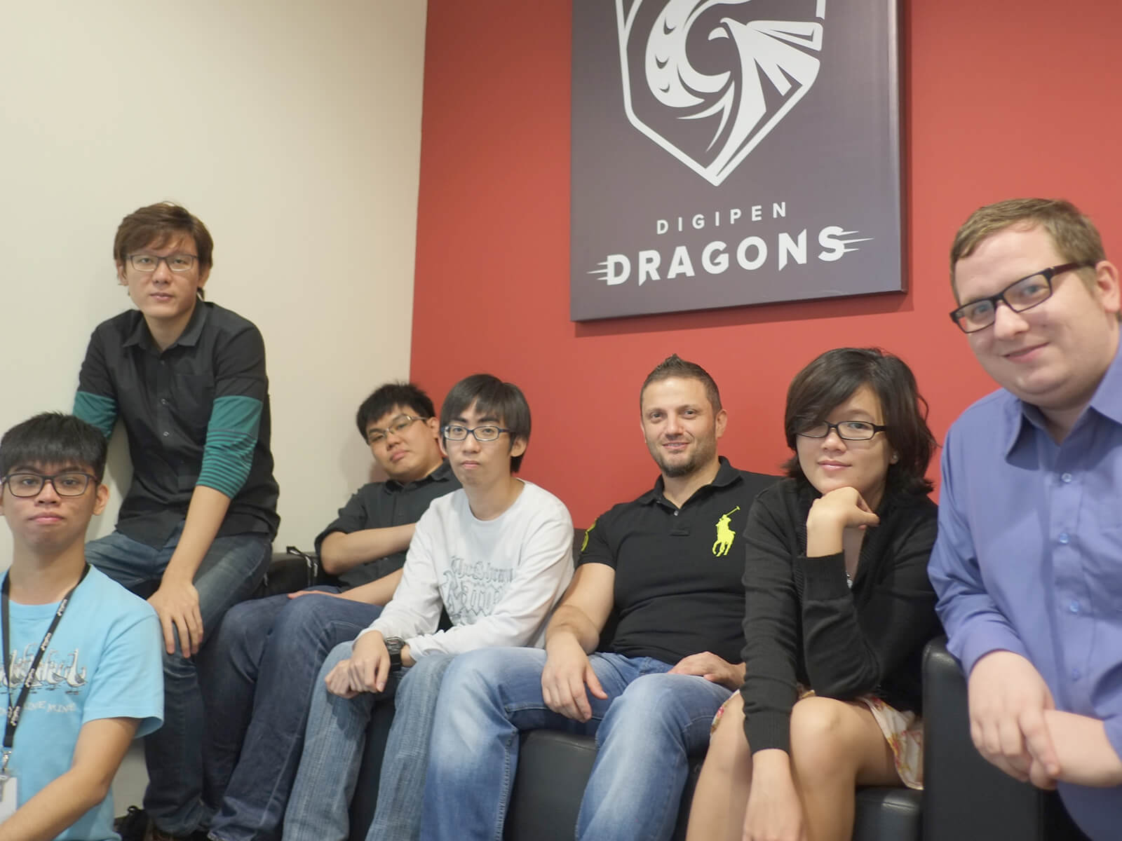 Members of the DigiPen (Singapore) Research and Development team sit at a coach below a poster of the DigiPen Dragons logo