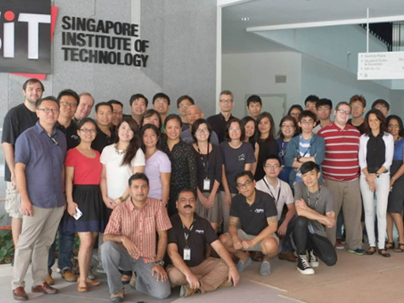 Dozens of DigiPen staff pose for a photo in front of a gray marble wall with the DigiPen and SIT logos