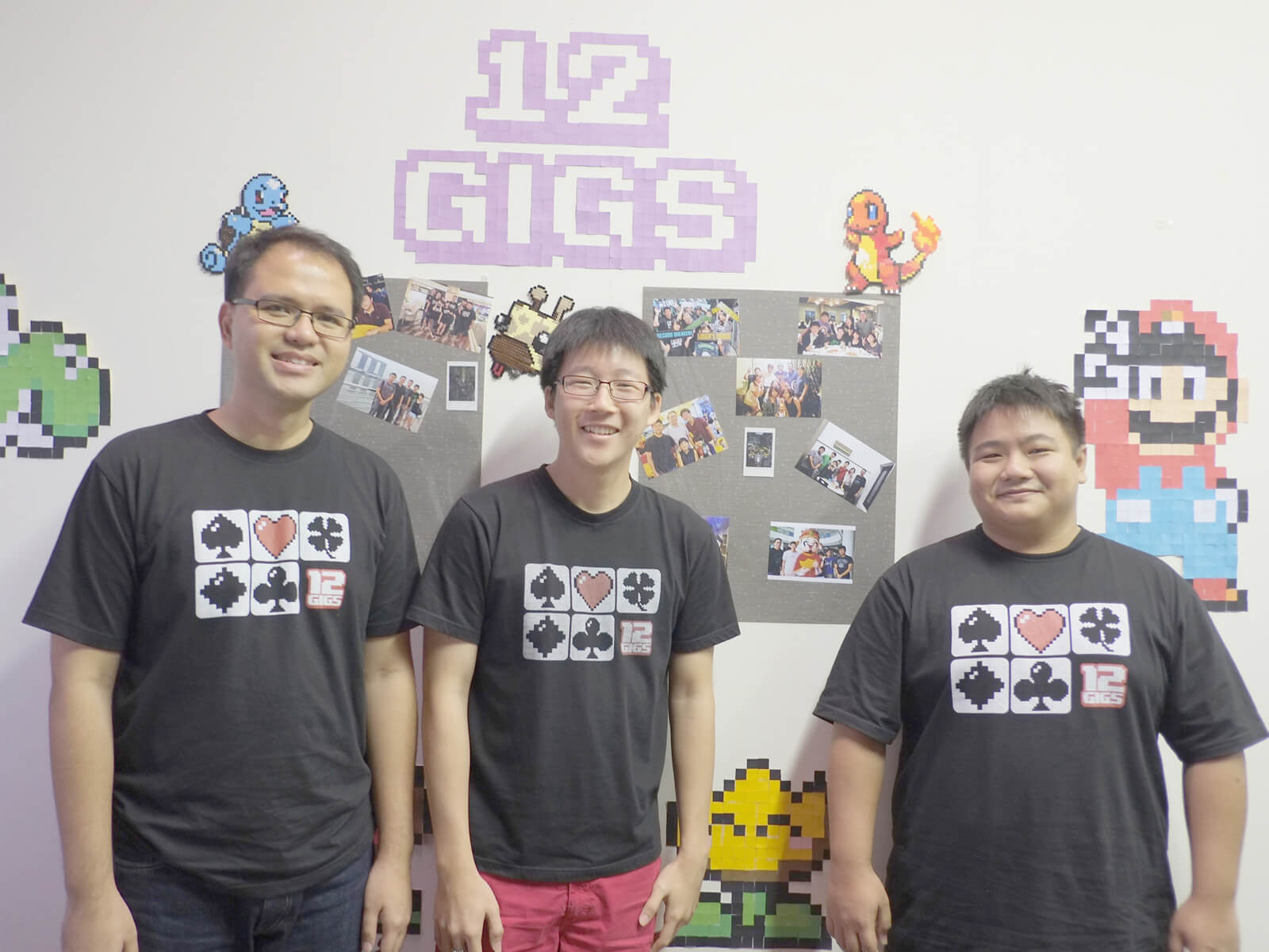 Three DigiPen graduates stand in front of a wall decorated with pixel art and the 12 Gigs logo wearing black t-shirts
