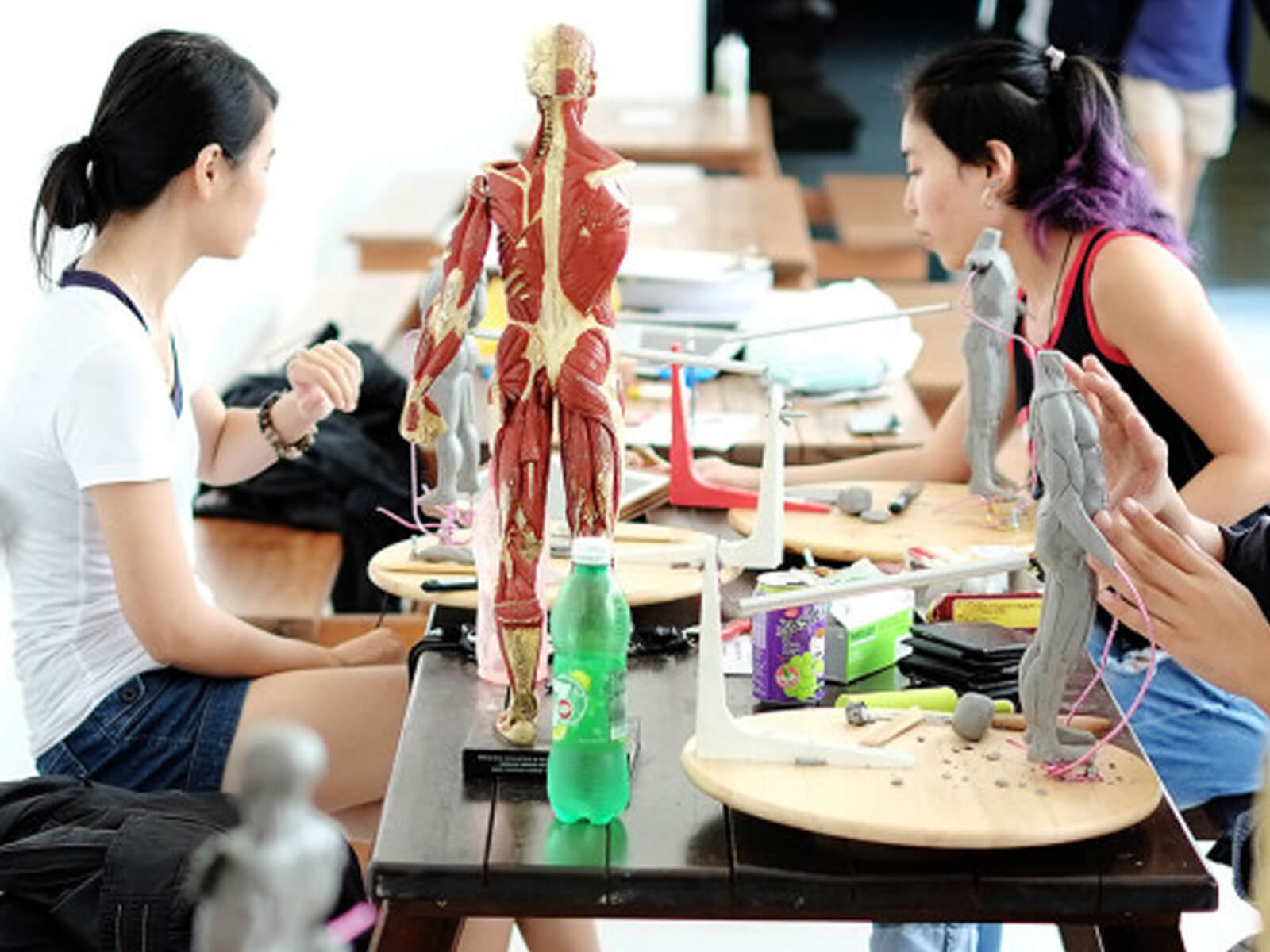 Students seated at a picnic table sculpt human figures out of clay. A plastic human musculoskeletal figure acts as a model.