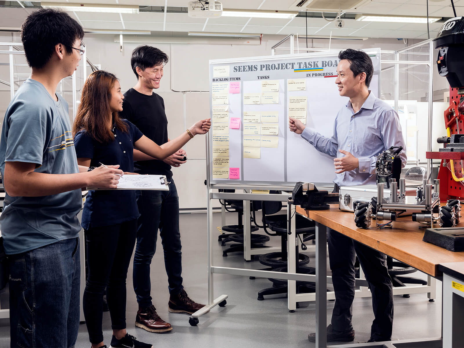 Three students and a teacher stand at a task board in a classroom lab.