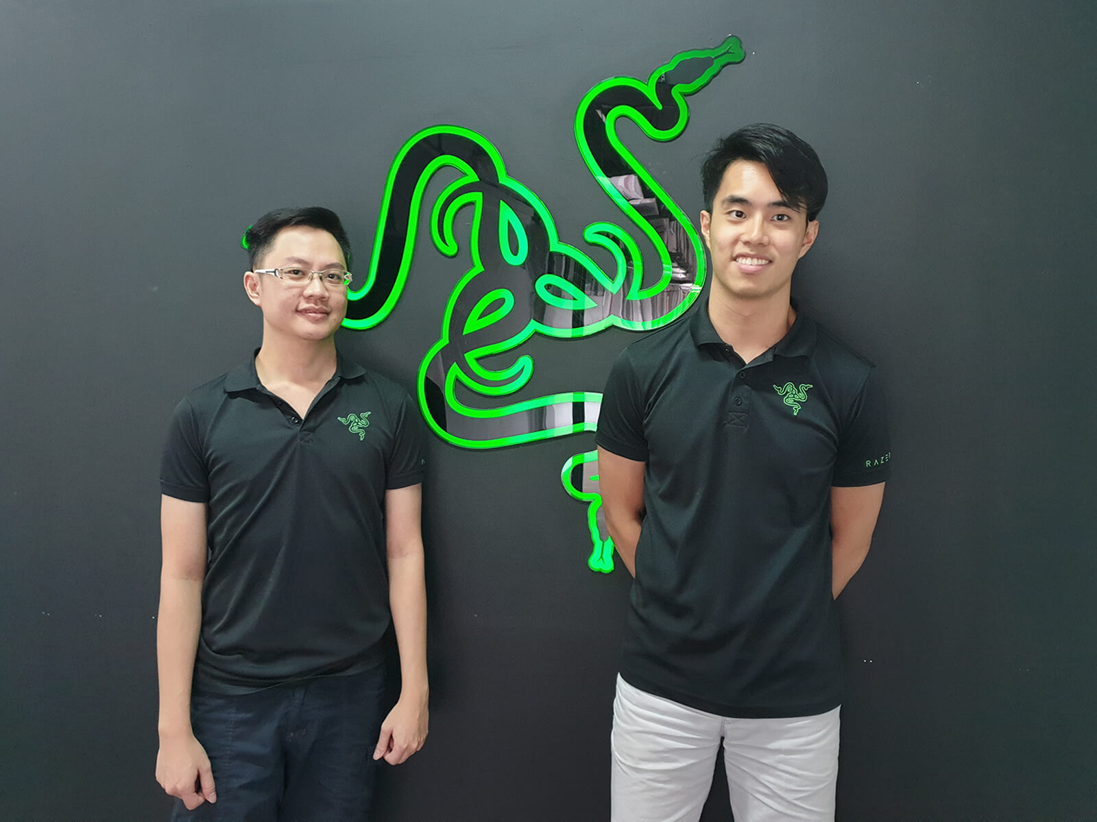 Two men wearing polo shirts with the green Razer logo stand by a gray wall, on either side of a much larger Razer logo.