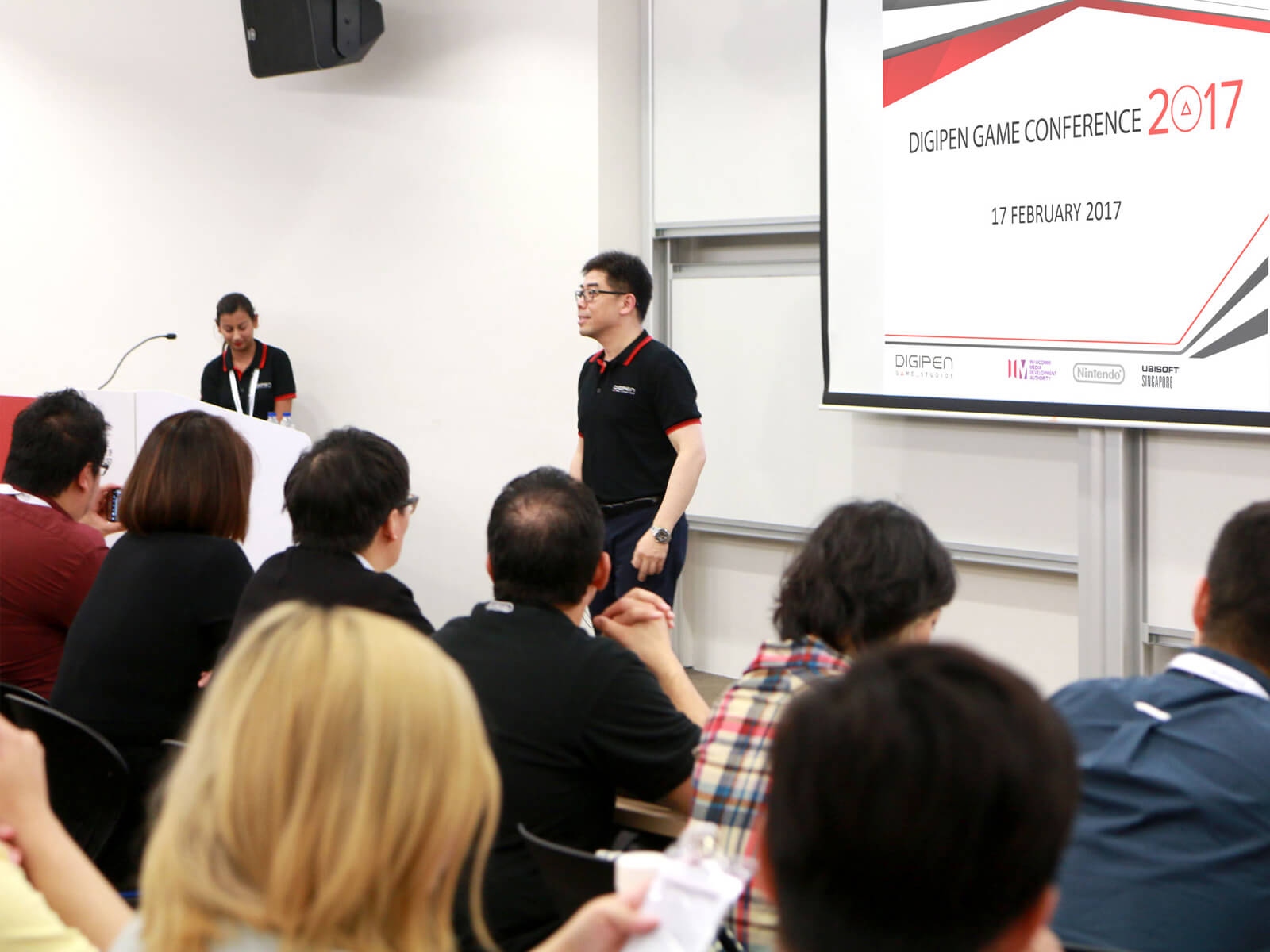 DigiPen COO Jason Chu addresses a room of industry professionals in a conference room. A projection screen is behind him.