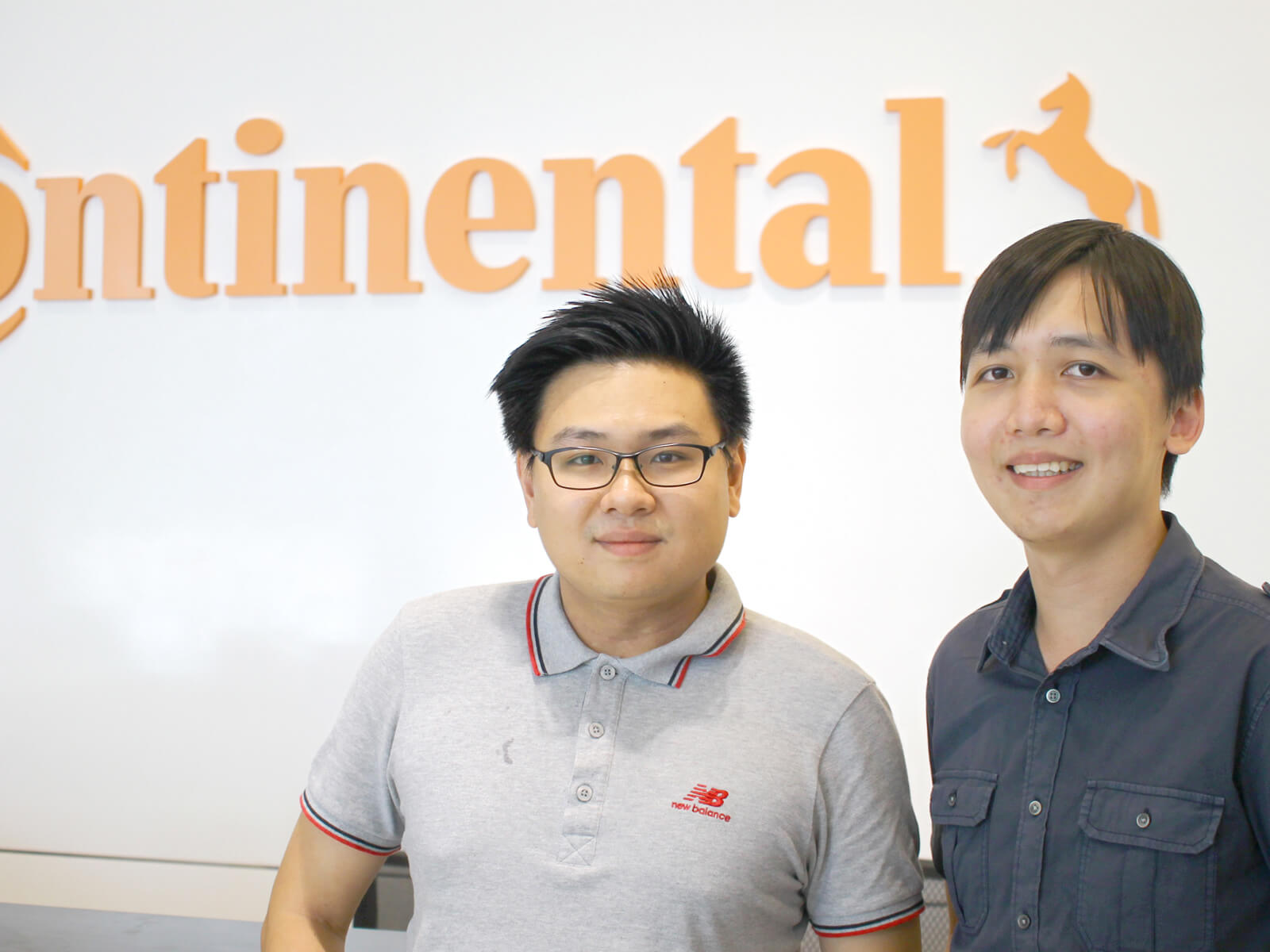 DigiPen (Singapore) alumni Jason Ngai and David Seah pose for a photo in front of a white wall adorned with the orange Continental logo