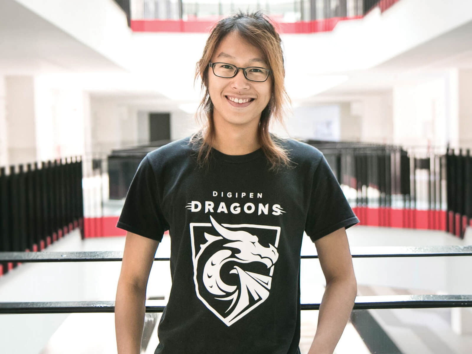 DigiPen graduate Jovi Kartolo poses for a photo at a railing on the second floor wearing a black DigiPen Dragons t-shirt