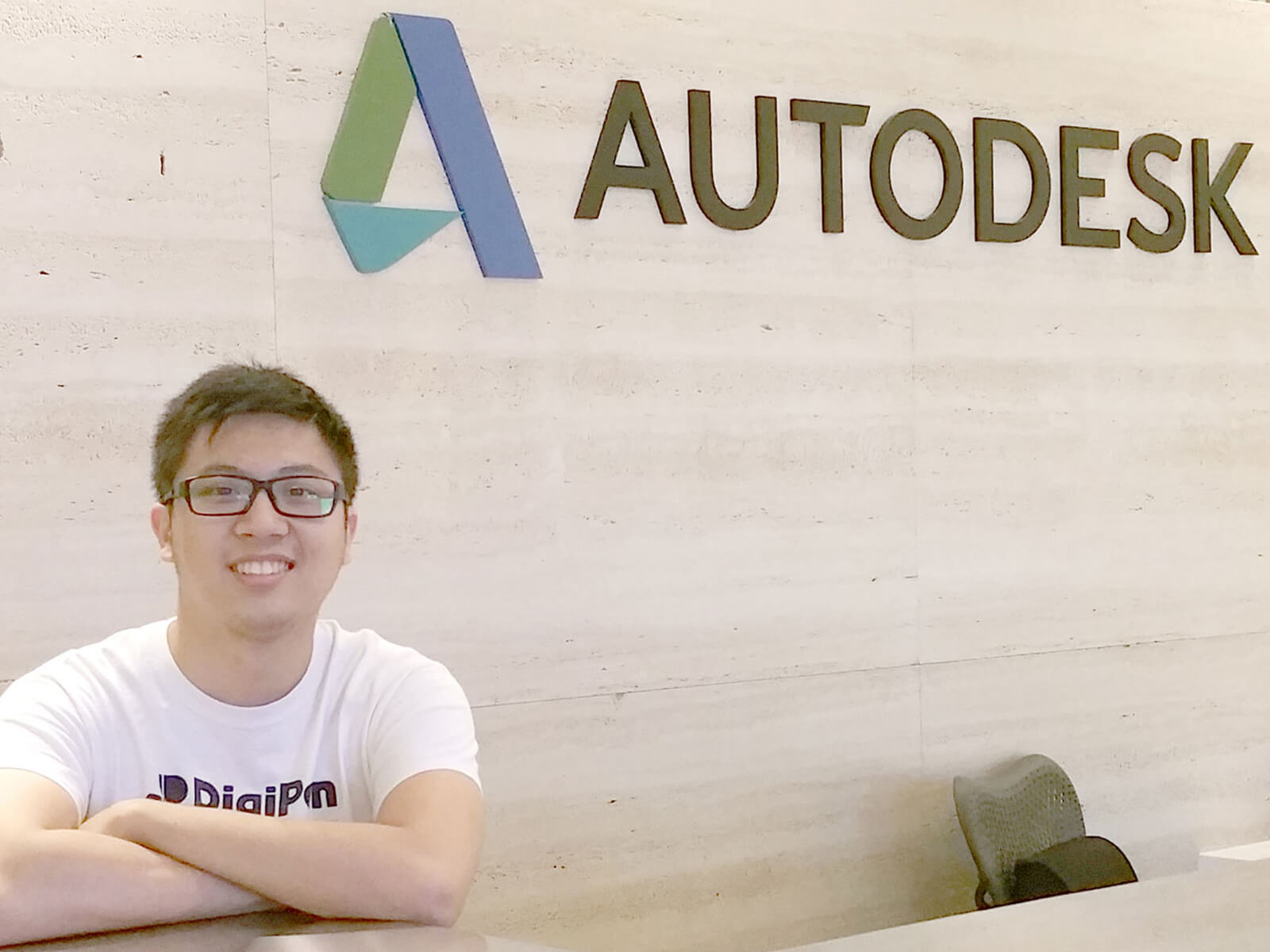 Graduate Julian Teh stands behind a counter with the Autodesk logo on a wall behind him