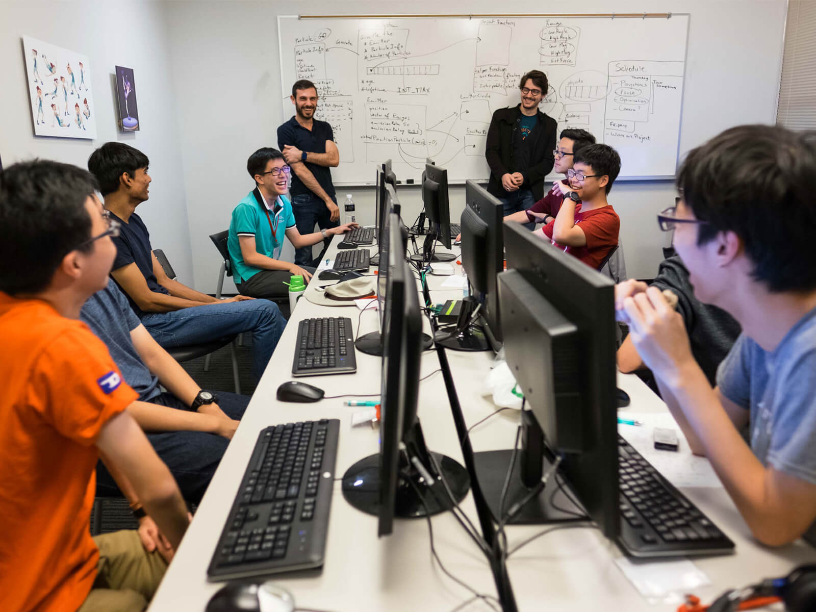 Several students sit at a long desk laughing as do their instructors, standing at a white board at the end of the table