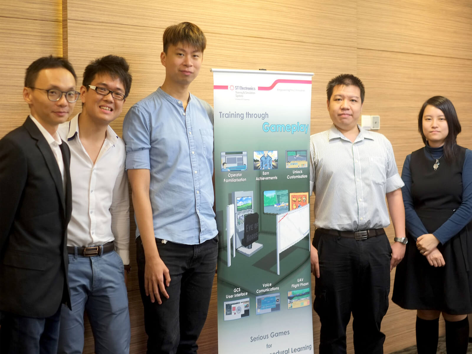 Team from ST Electronics poses for a photo with a vertical banner explaining a learning initiative using gameplay mechanics